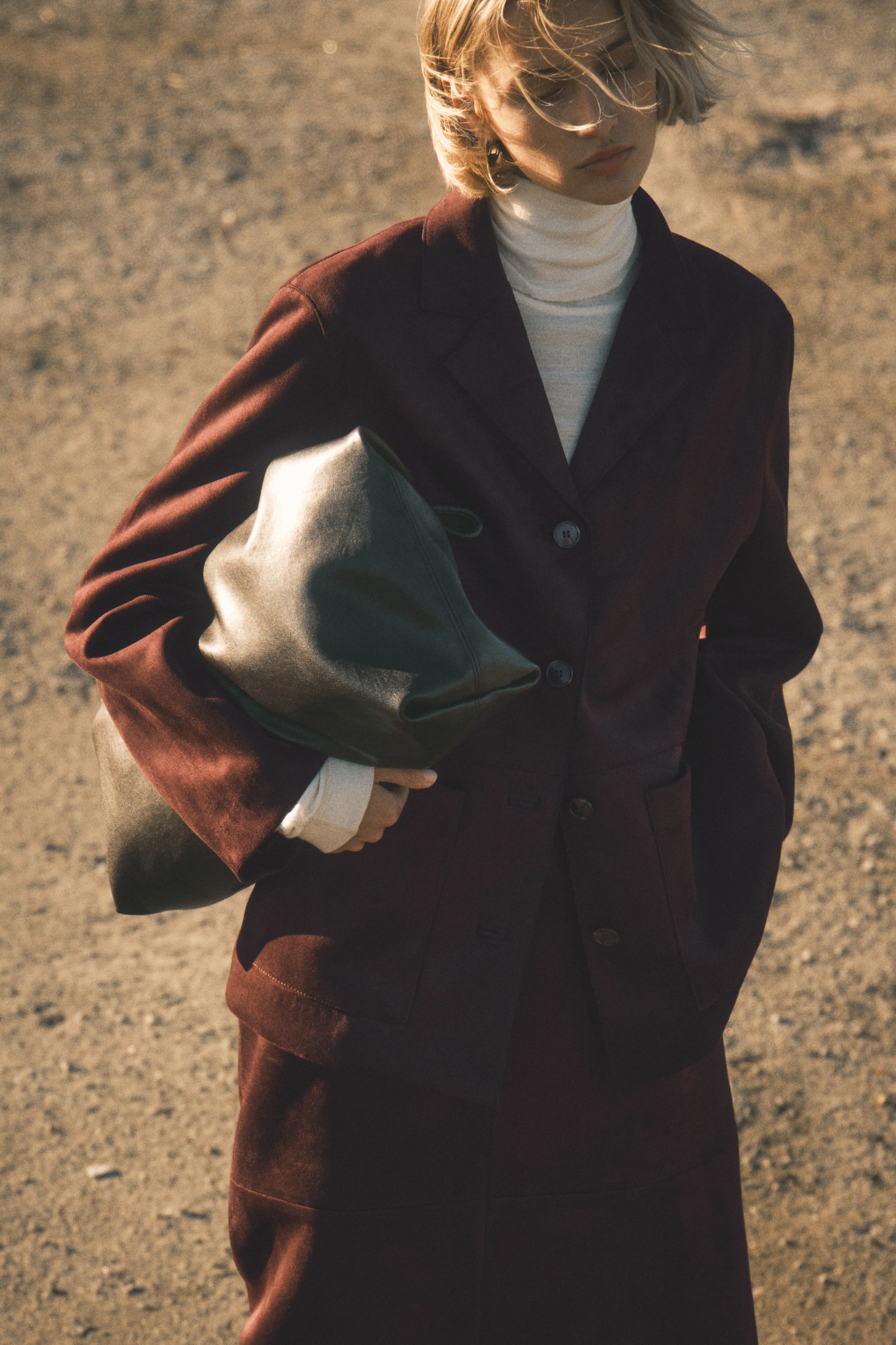 查看大圖: 一件深紅色的單排扣西裝外套,設有兩顆扣子、缺角領和翻蓋口袋,從略微傾斜的正面視角穿戴在一件米色羅紋高領毛衣和一條深紅色裙子上。一個柔軟的深綠色皮革手提包被夾在腋下。