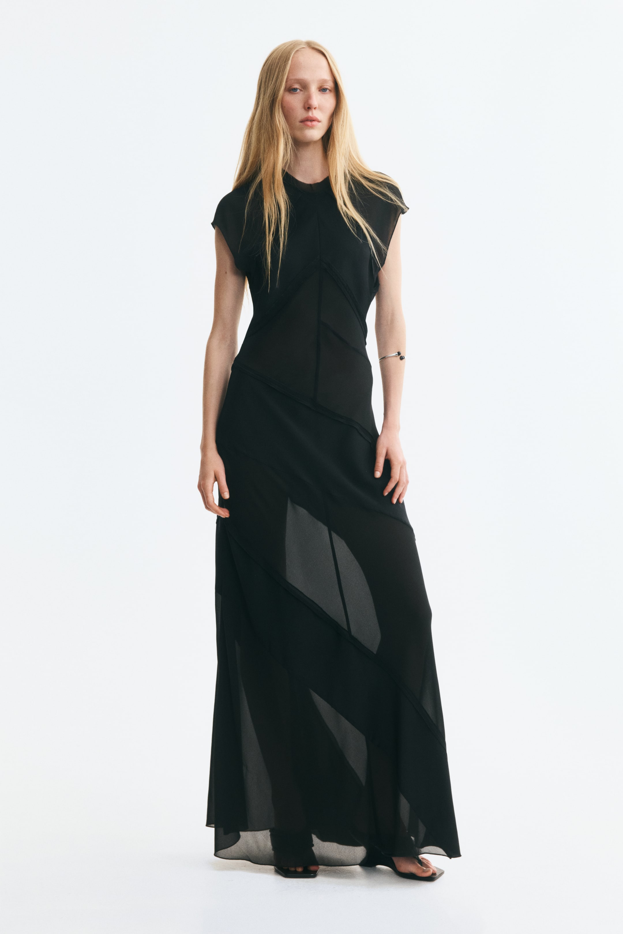 View larger image: The model is wearing a black fitted maxi dress featuring diagonal, inside-out-look seams, a round neckline, and cap sleeves. The dress combines structured woven fabric with sheer chiffon panels, creating a semi-transparent layered effect on the skirt.
