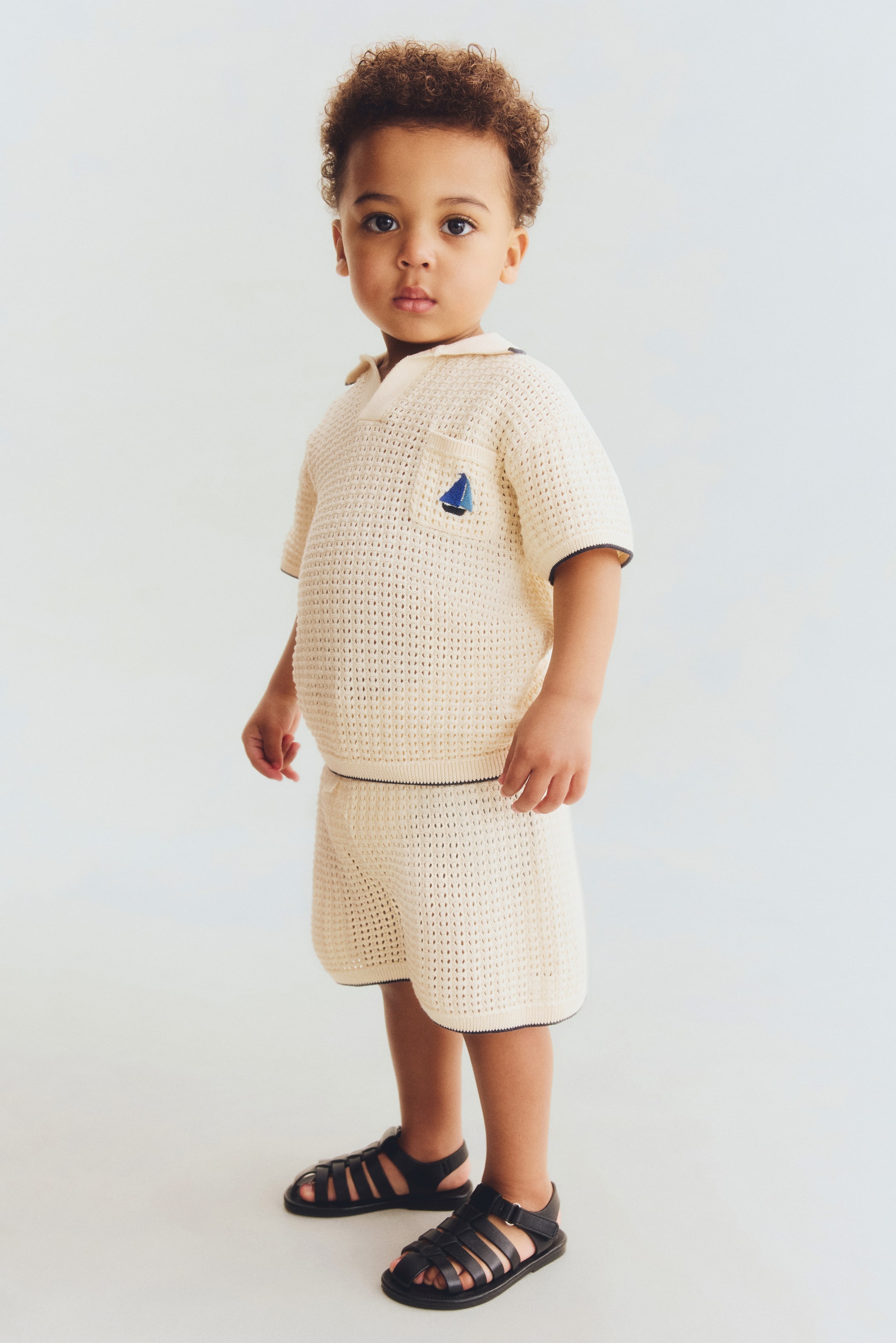 Grotere afbeelding bekijken: Een crème opengewerkt gebreide polo met korte mouwen en bijpassende shorts worden gedragen, met donkere biezen op de kraag, knopenlijst, mouwboorden en zoom, en een blauw zeilbootborduursel op het borstzakje van de top. Zwarte sandalen met bandjes maken de look compleet.