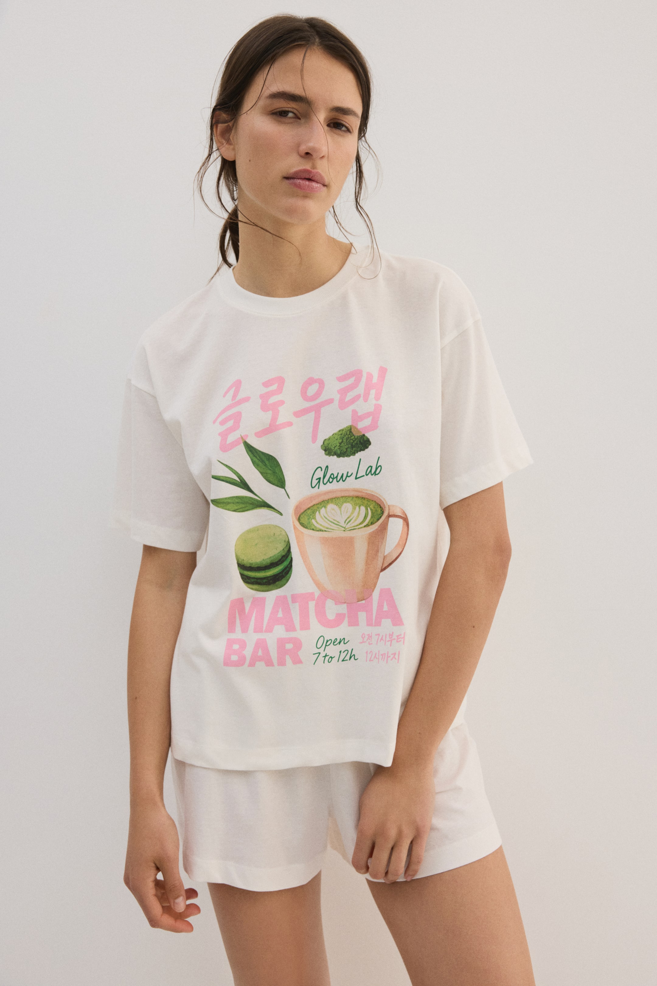 View larger image: The white, relaxed-fit, short-sleeved t-shirt showcases a vibrant graphic print on its front. This print features a matcha latte in a peach-colored mug, a green macaron, matcha powder, green leaves, and text including "Glow Lab" and "Matcha Bar" in pink, alongside Korean text.