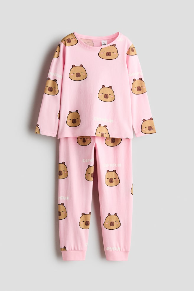 Ein rosa Pyjama-Set hängt nach vorne gerichtet und besteht aus einem langärmeligen Oberteil mit Rundhalsausschnitt und passenden langen Hosen. Beide Kleidungsstücke zeigen ein sich wiederholendes Muster von Cartoon-Capybara-Gesichtern und dem Text "CAPYFUN", wobei die Hosen mit elastischen Bündchen an den Knöcheln gestaltet sind. - 1