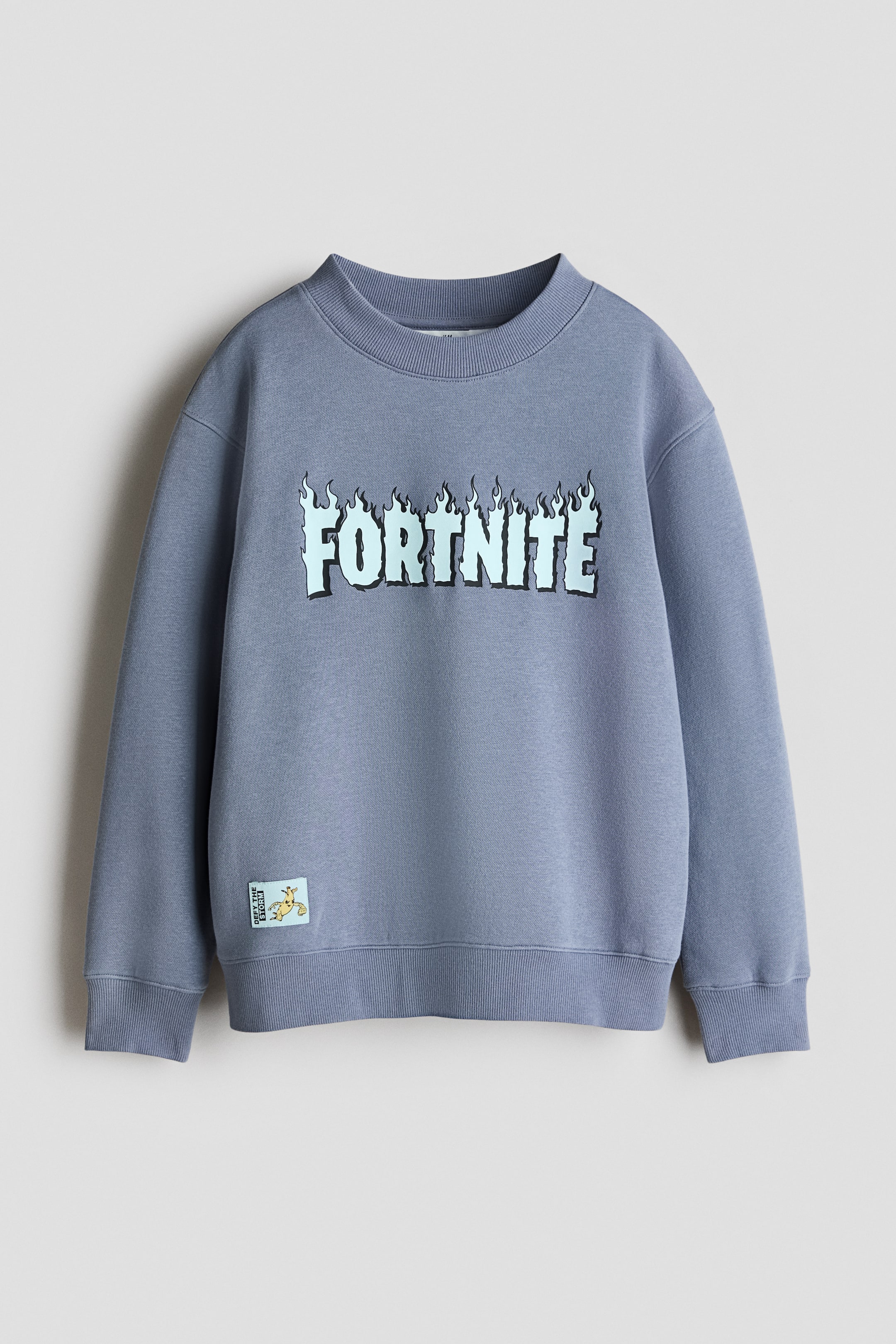 View larger image: A gray-blue crewneck sweatshirt is shown from the front, featuring a light blue "Fortnite" logo with a flame design across the chest. Ribbed cuffs and a ribbed hem are visible, and a small character patch adorns the lower left.