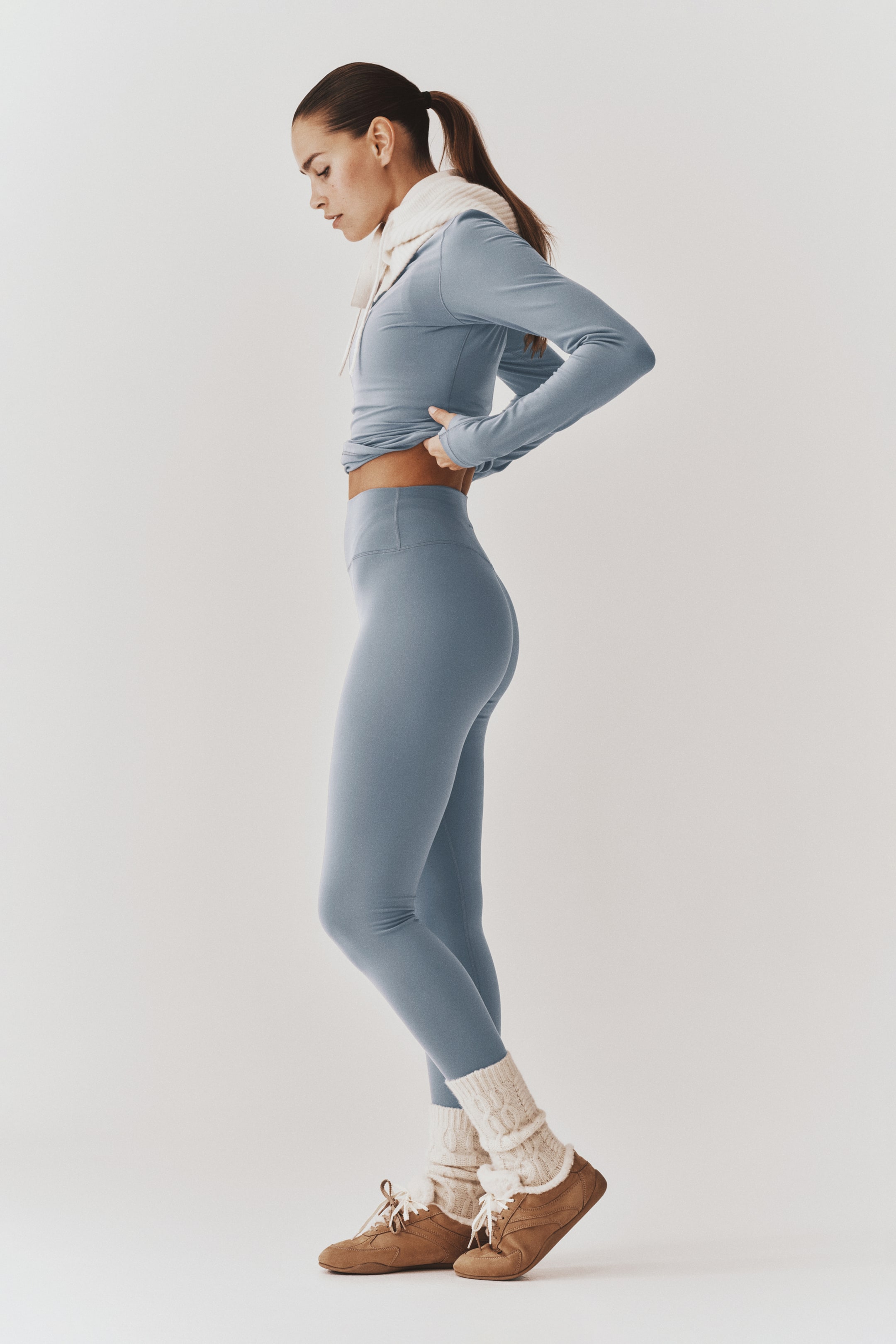Büyük resmi görüntüle: Tozlu mavi bir activewear seti, vücuda oturan uzun kollu bir üst ve yüksek belli tayttan oluşuyor; model tarafından yandan profil görünümünde giyilmiş. Model, üstün etek ucunu ayarlayarak taytın yüksek bel bandını öne çıkarıyor; kombin, krem rengi slouch çoraplar ve kahverengi bağcıklı ayakkabılarla tamamlanmış.