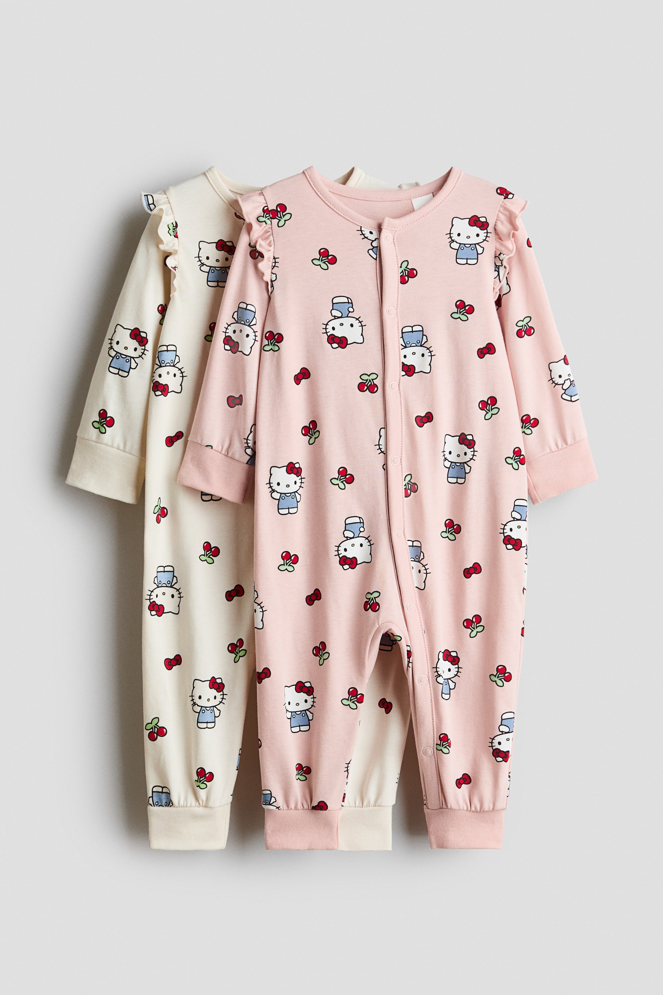 Agrandir l'image: Deux combinaisons de nuit en jersey de coton, l’une rose clair et l’autre crème, sont suspendues de face et présentent un imprimé intégral de personnages de dessin animé, de nœuds et de cerises. Les deux combinaisons de nuit sont dotées d’encolures rondes, de détails volantés aux épaules, de fermetures à pression descendant sur le devant et sur une jambe, ainsi que de larges bords-côtes aux poignets et aux chevilles.