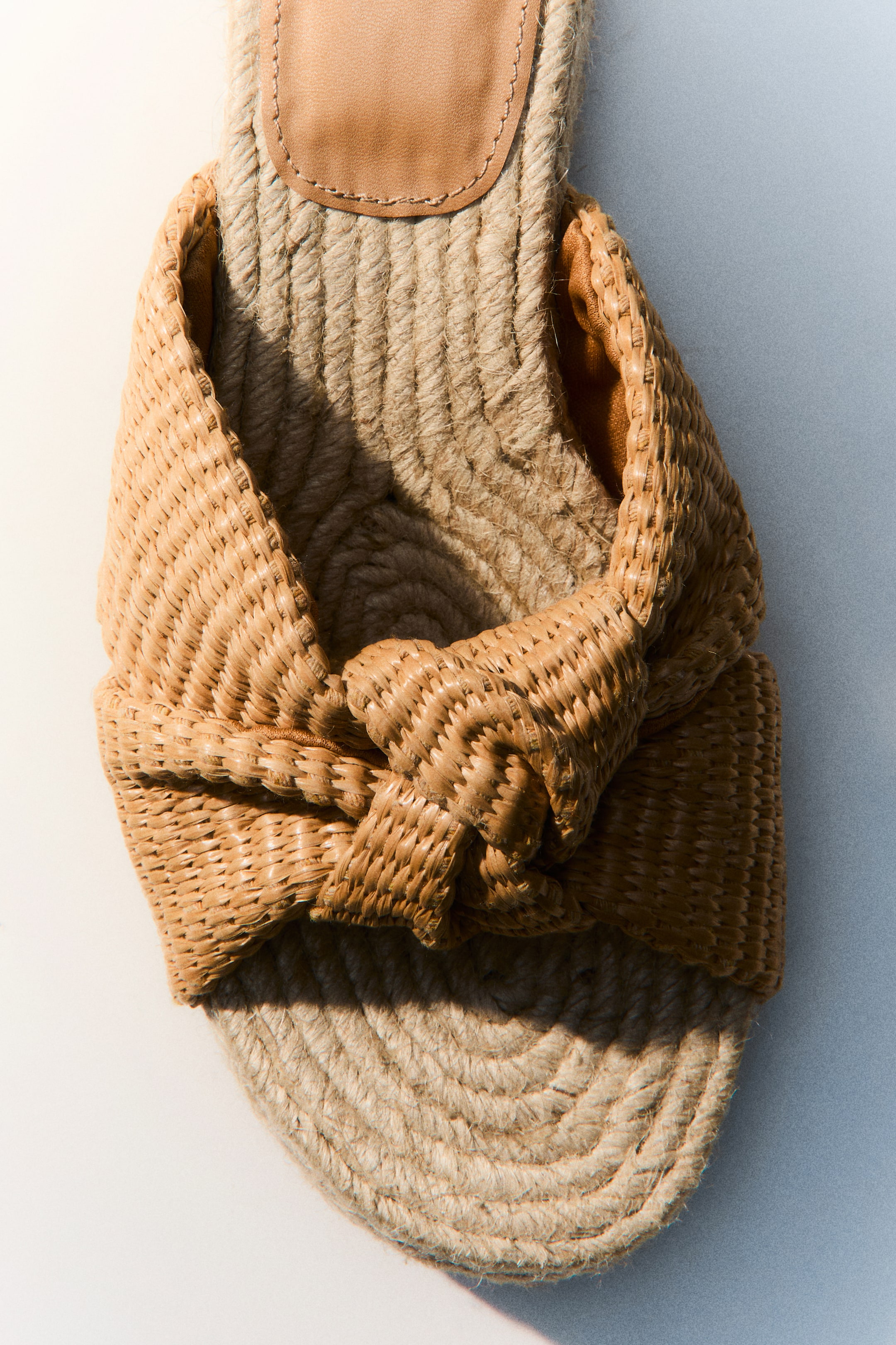 Visualizza immagine più grande: Una ciabatta espadrillas color cuoio mette in mostra la sua suola in corda di juta e una tomaia ampia in rafia intrecciata. La tomaia in rafia forma un design annodato sulla parte anteriore, mentre una piccola patch in pelle color cuoio è visibile sulla soletta.
