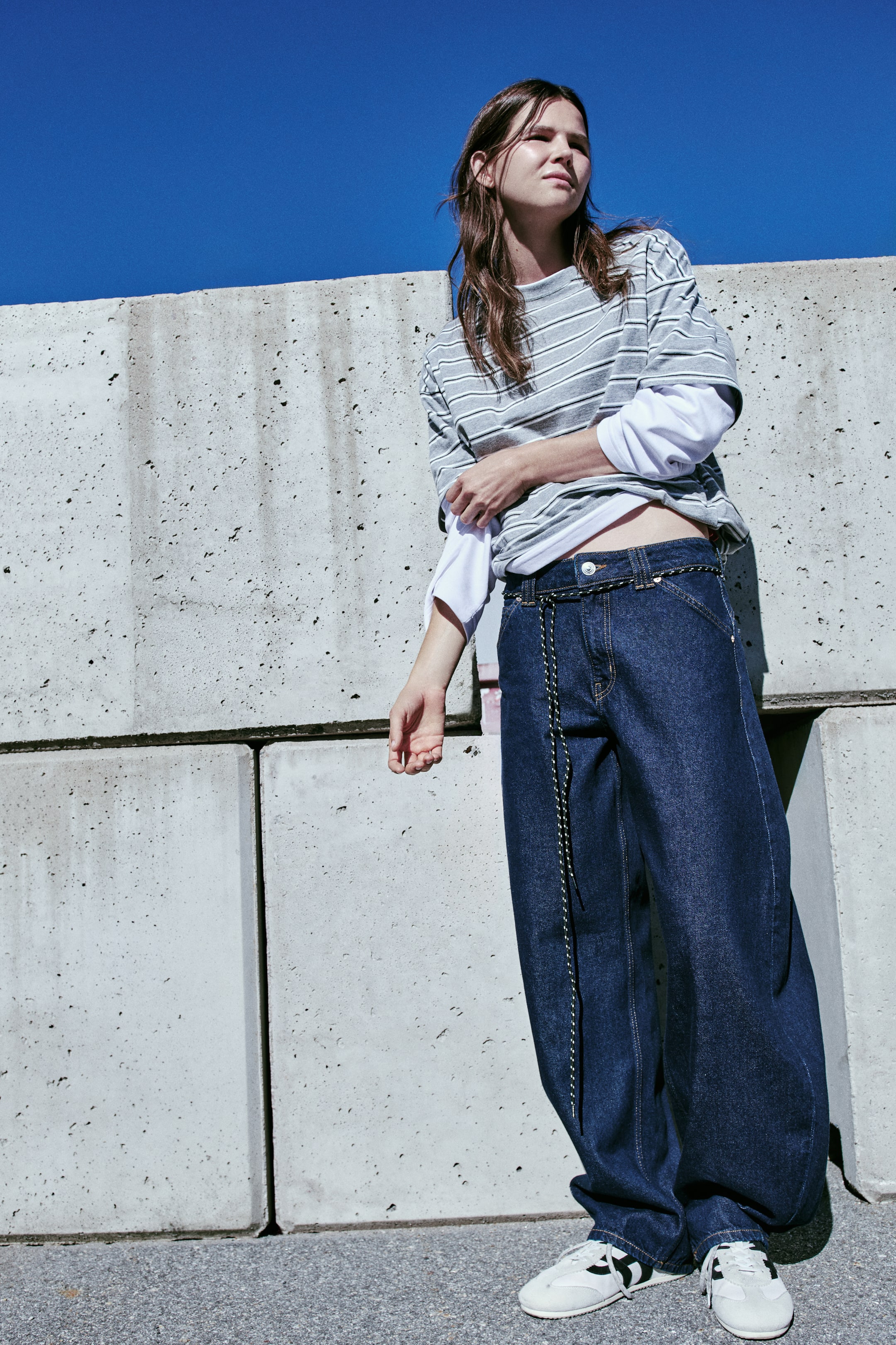 View larger image: A pair of dark blue denim jeans is shown from the front, featuring a regular waist with a button fastening, a thin cord belt, and classic five-pocket styling. The jeans have a relaxed fit with voluminous, curved legs that taper towards the hems, finishing at the feet with minimal stacking over white trainers.