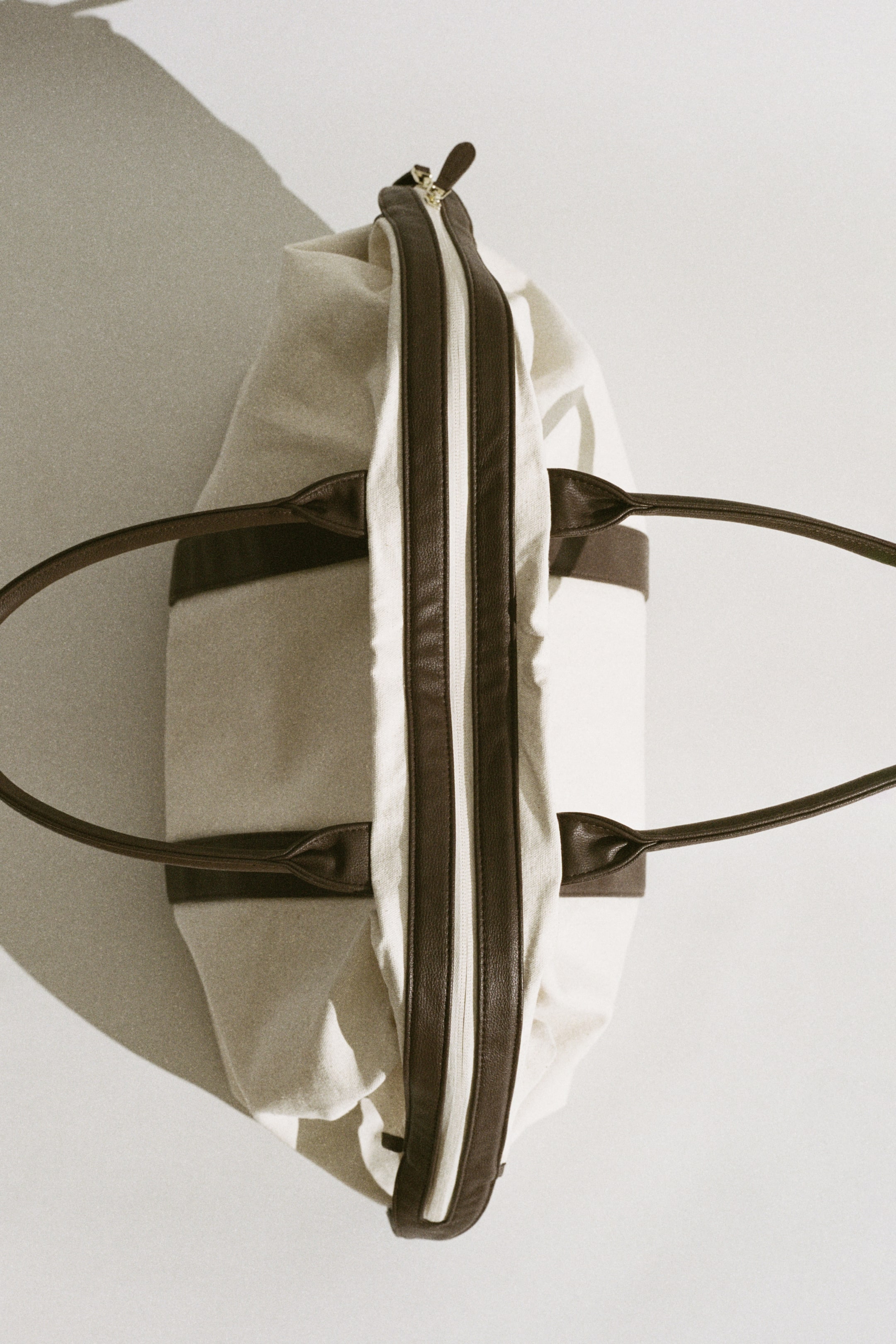 Vis større bilde: En duffelbag er utstyrt med en off-white lerretskropp med mørkebrune læraktige stropper og detaljer, med en gullfarget glidelås langs den litt åpne toppen.