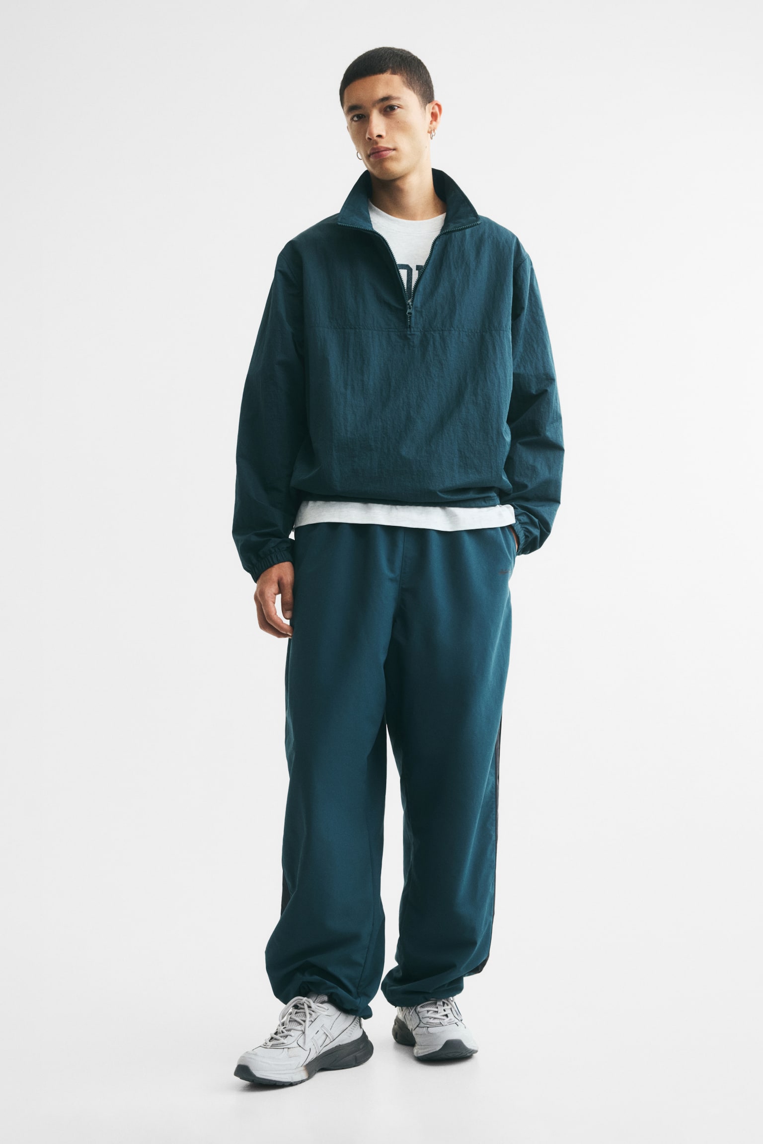 Dark teal track pants, laid flat, are featuring covered elastication at the waist and contrasting black stripes with piping running down each side. Elasticated drawstrings with cord stoppers are visible at the hems, presenting a loose fit. - 1