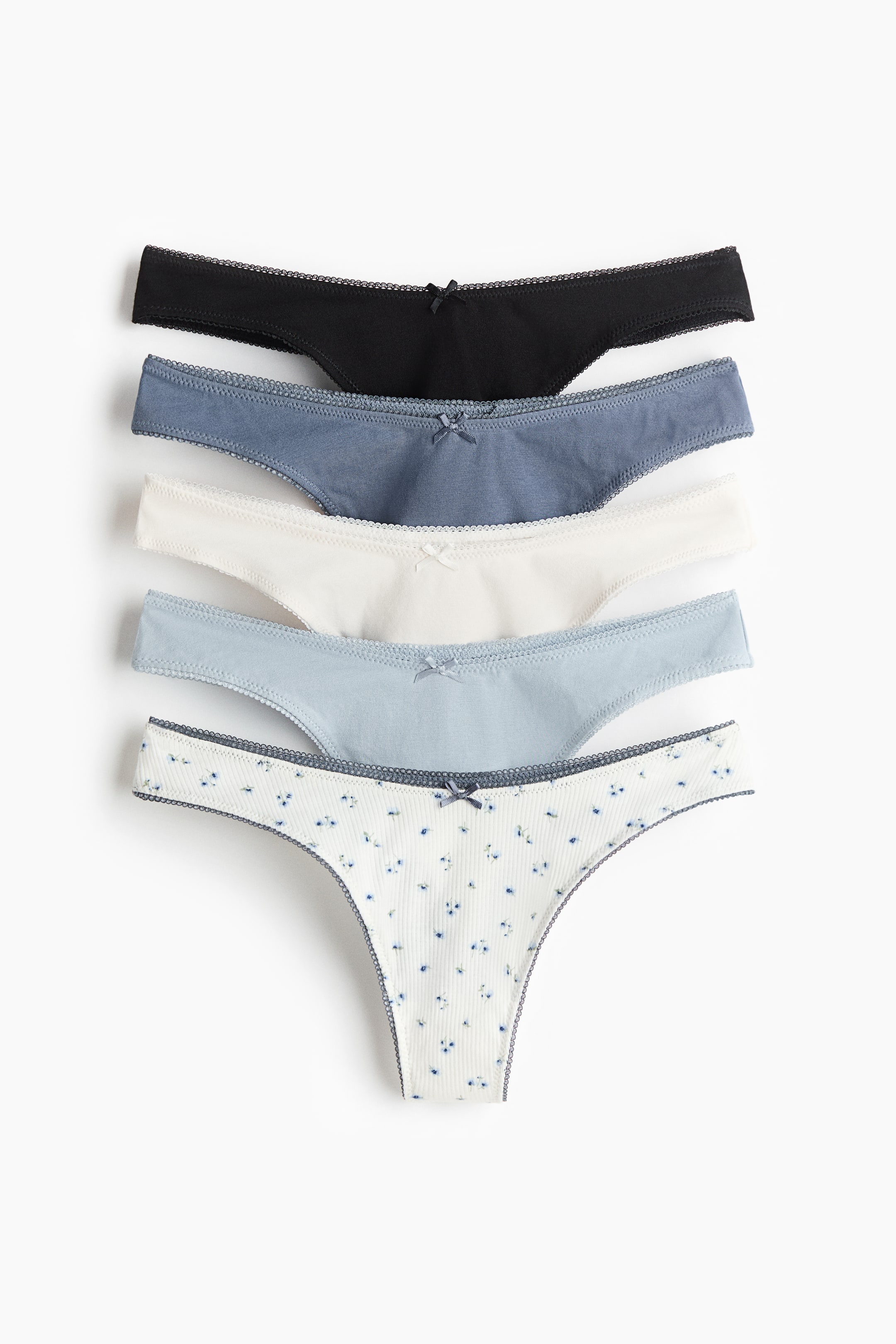 Visualizza immagine più grande: Un set di cinque perizomi in jersey di cotone con vita bassa a V e cavallo foderato mostra la parte frontale. Ciascun perizoma presenta un piccolo fiocco al centro davanti e una rifinitura picot lungo la vita e gli orli delle gambe, proposti nei colori nero, blu polvere, bianco, azzurro e in una variante bianca a costine con motivo floreale blu.
