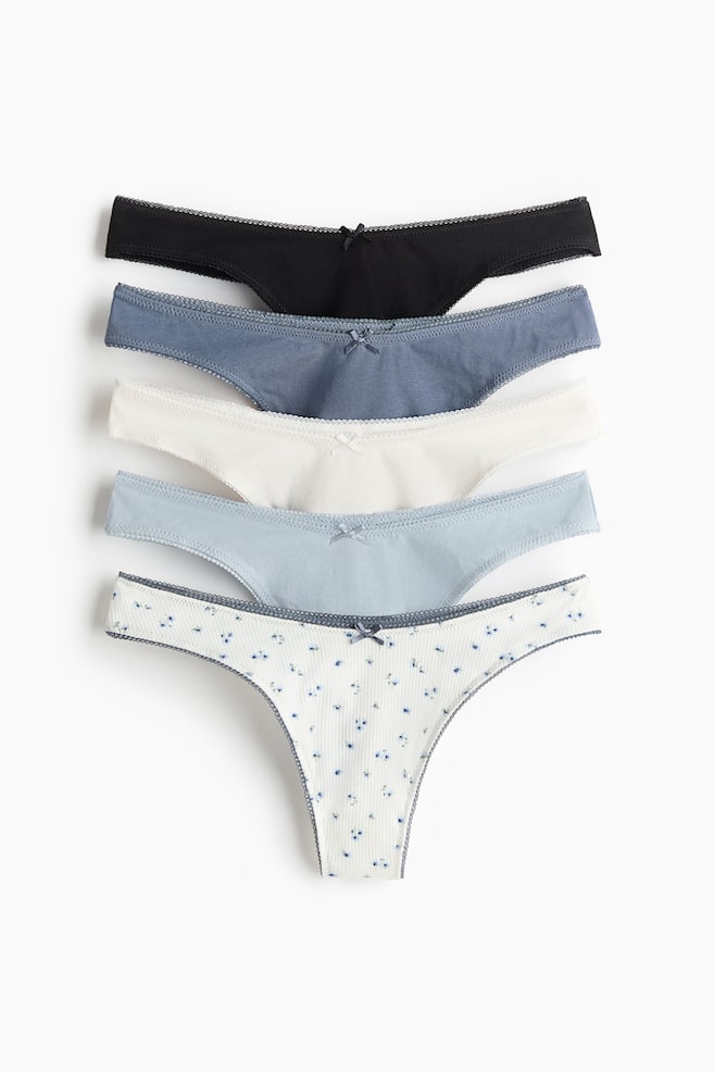 A set of five thong briefs in cotton jersey with a low V-shaped waist and a lined gusset, shown from the front. Each brief features a small bow at the centre front and picot trim along the waist and leg openings, in black, dusty blue, white, light blue and a white ribbed pair with a blue floral pattern. - 1