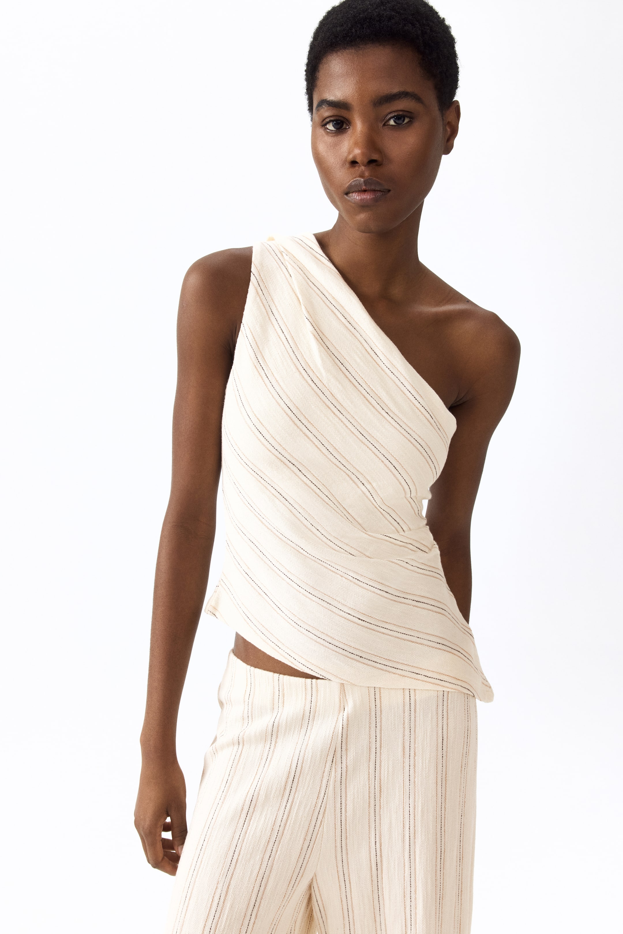 Größeres Bild anzeigen: Ein cremefarbenes One-Shoulder-Top und ein dazu passendes Hosen-Set, mit dünnen braunen und schwarzen Streifen sowie einem strukturierten Stoff, wird aus einem leicht frontalen linken Winkel präsentiert. Das Top ist mit einer asymmetrischen Drapierung gestaltet, und die Hose ist mit vertikalen Streifen versehen.