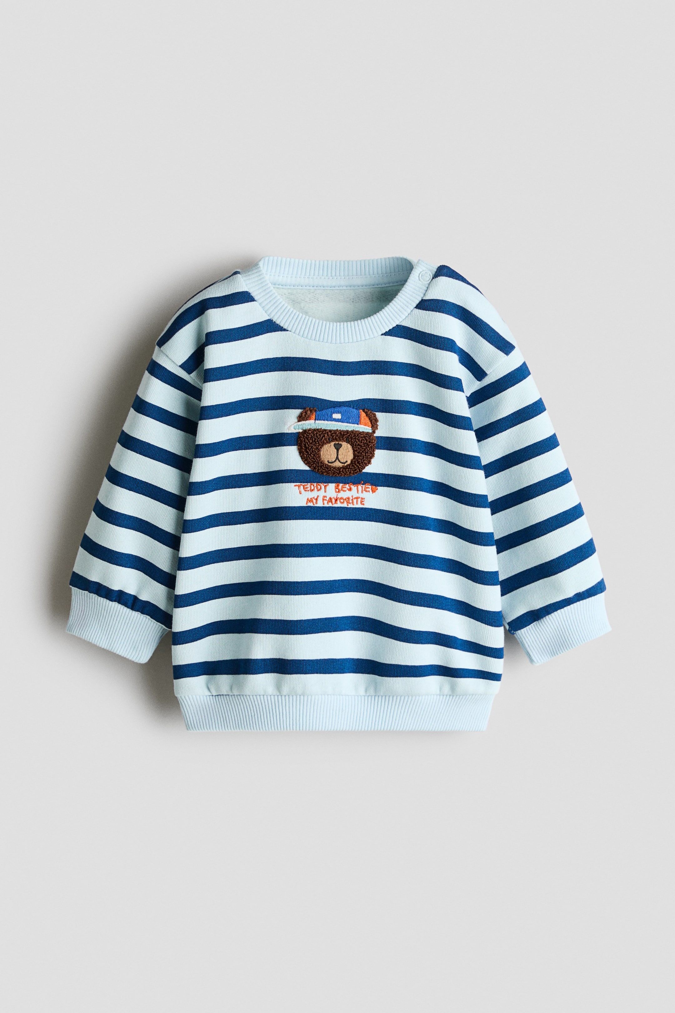Visa större bild: En ljusblå och mörkblå randig sweatshirt visas framifrån, med en ribbad rundhals med axelknäppning, ribbade muddar och ribbad nederkant. På mitten av bröstet visas en broderad brun teddybjörn med en blå keps, placerad ovanför orange broderad text som lyder "TEDDY BESTIES MY FAVORITE".