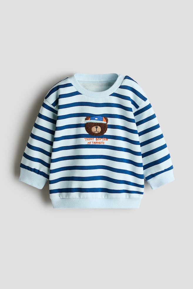 Ein hellblau und dunkelblau gestreifter Sweatshirt wird in der Vorderansicht gezeigt und verfügt über einen gerippten Rundhalsausschnitt mit Schulter-Druckknopfverschluss, gerippte Bündchen und einen gerippten Saum. Auf der Brustmitte ist ein braun gestickter Teddybär mit blauer Kappe zu sehen, platziert über der orangefarbenen gestickten Aufschrift âTEDDY BESTIES MY FAVORITEâ. - 1