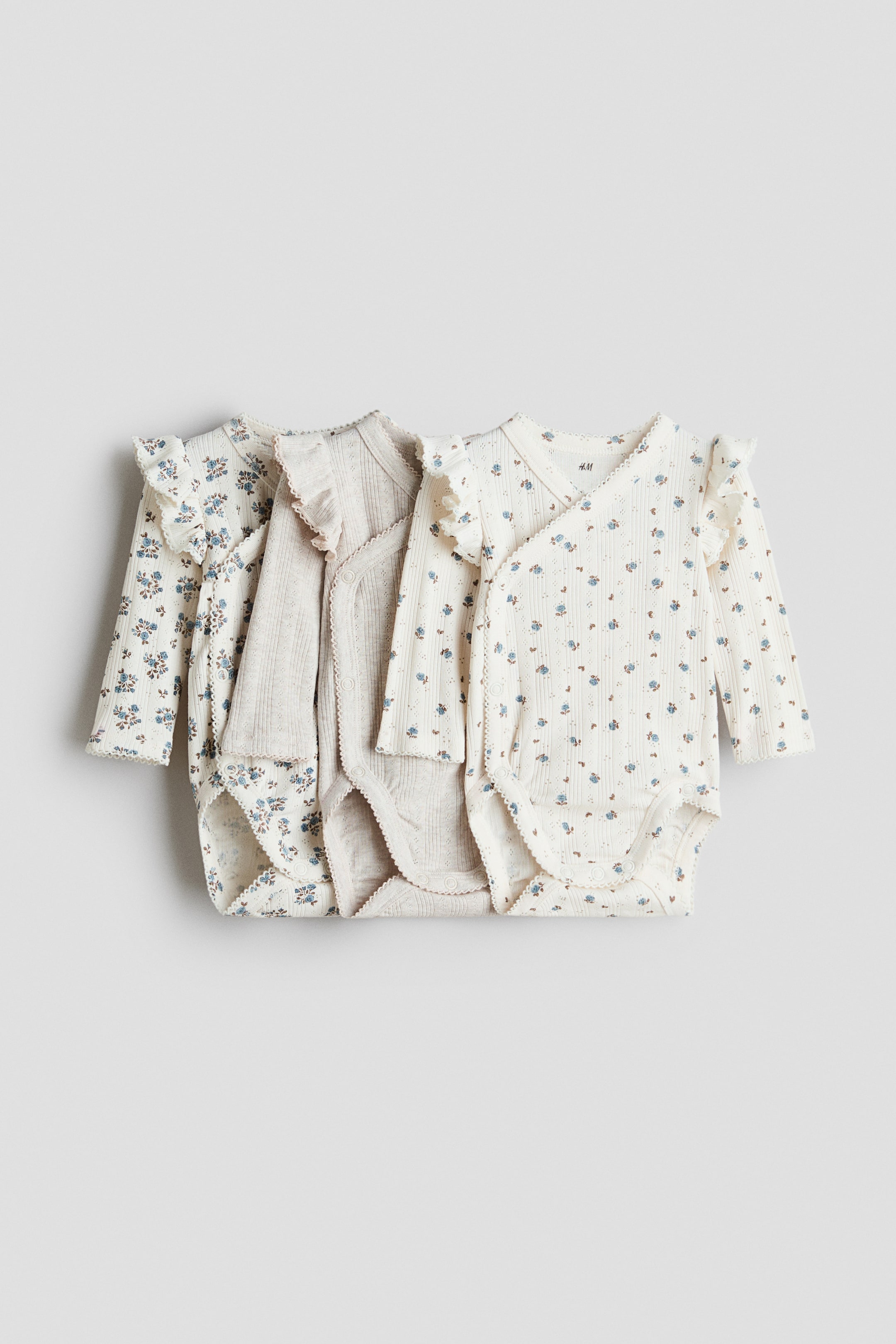 Visa större bild: Långärmade bodysuits visas framifrån, med en omlottdesign med delikata picotkanter och volanger över axlarna. Två bodysuits visar ett litet blått blommönster på krämfärgad bomullspointelle jersey, och en är en enfärgad beige bomullspointelle jersey, alla med tryckknappar på sidorna och i botten.