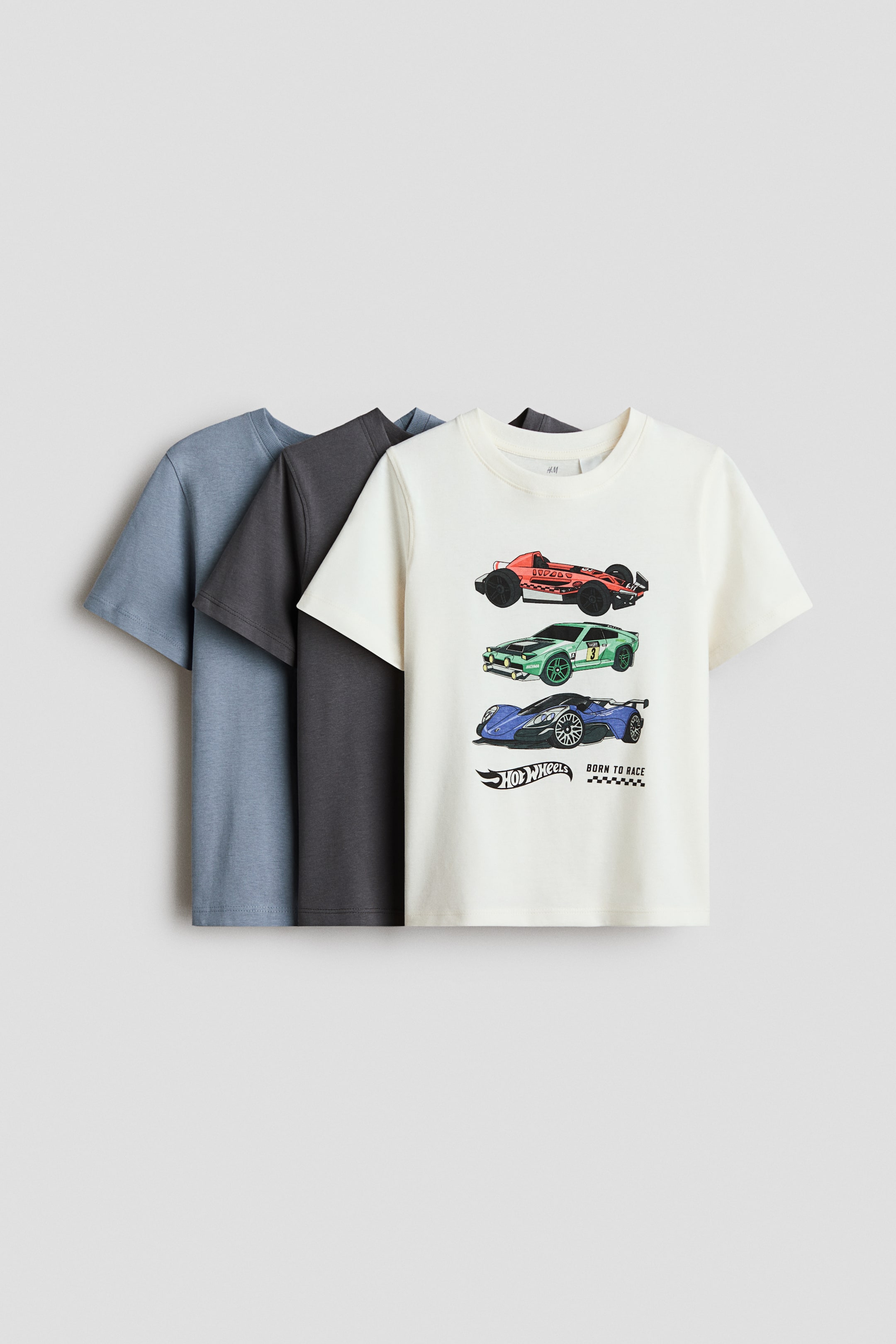 Se større billede: Tre kortærmede t-shirts er stablet, let overlappende, med deres forsider vendt fremad. Den forreste t-shirt er off-white og viser et grafisk print af tre racerbiler samt et "Hot Wheels" logo, mens en mørkegrå t-shirt og en lyseblå t-shirt er synlige bagved. Alle tre t-shirts har en ribkantet rund halsudskæring.