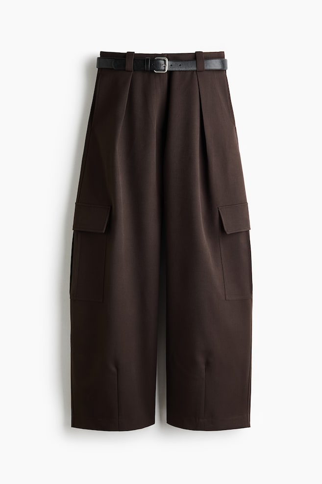 This pair of dark brown, wide-leg trousers is shown from the front, featuring a high waist styled with a slim black belt with a silver buckle. The trousers feature front pleats, side cargo pockets with flaps, and small vertical slit details at the front of the hems. - 1