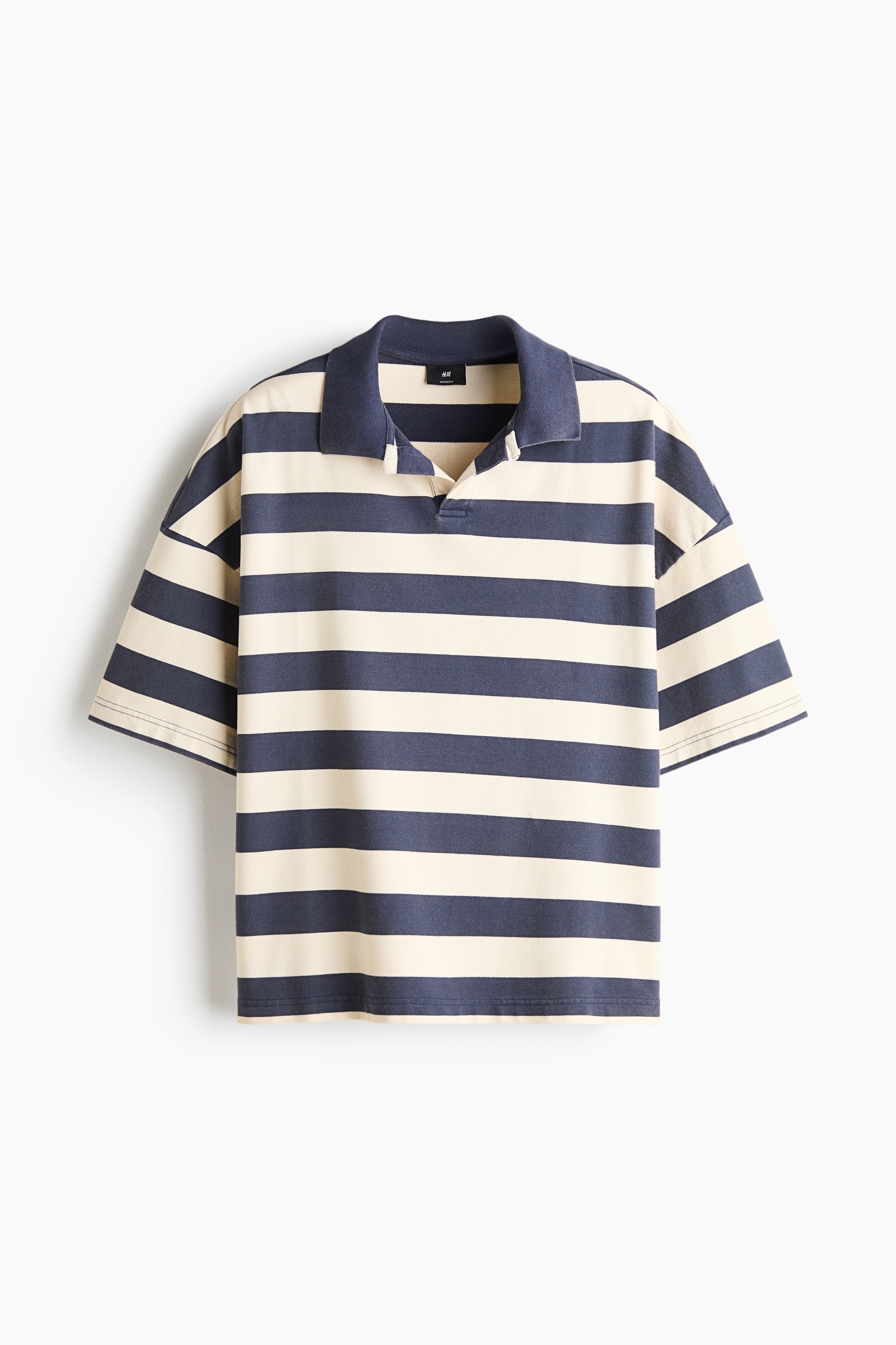 Een oversized poloshirt met korte mouwen en horizontale strepen in blauw en crÃ¨me wordt gedragen door het model. Het shirt, ontworpen met een open kraag en verlaagde schouders, wordt gecombineerd met een wit T-shirt en donkere wijde broek, terwijl een witte jas in de hand wordt gehouden.