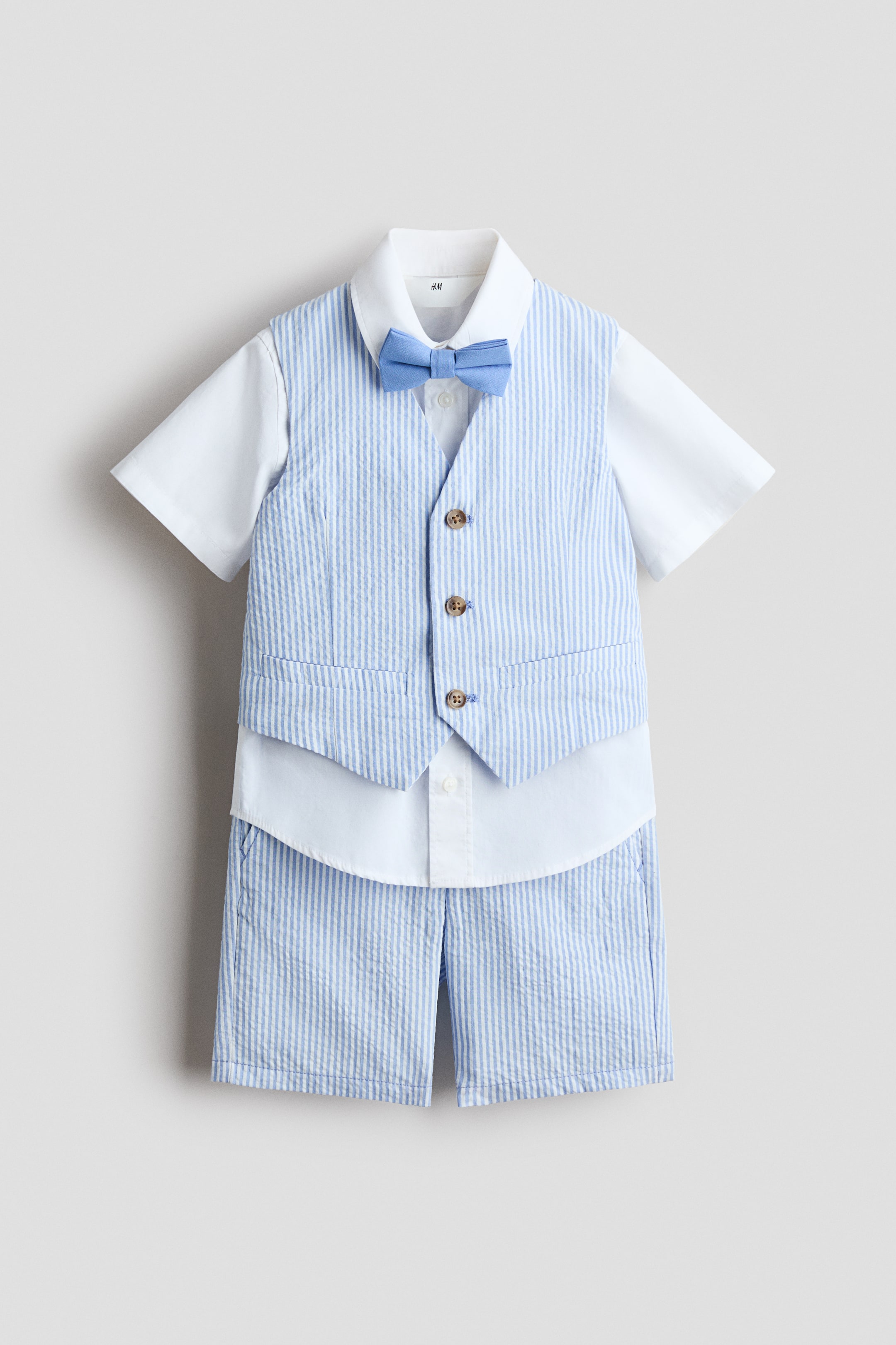 View larger image: A children's four-piece set is displayed front-facing, featuring a white short-sleeved shirt with a turn-down collar, layered under a light blue and white seersucker-striped vest with visible buttons and faux welt pockets. Matching light blue and white seersucker-striped knee-length shorts with discreet side pockets are visible below, completed by a light blue bow tie at the shirt's collar.