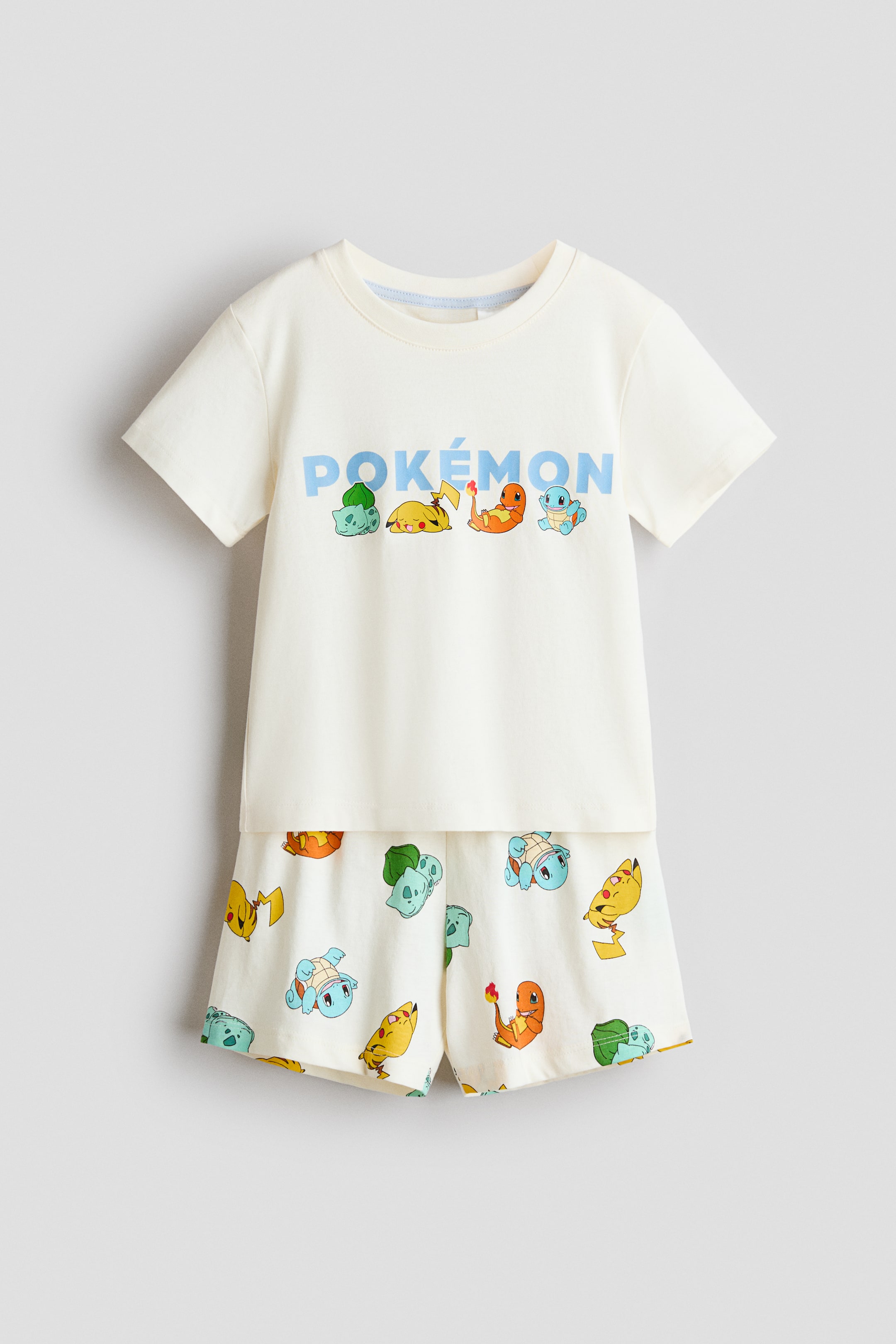 View larger image: A cream-coloured two-piece pyjama set is shown from the front, comprising a short-sleeved t-shirt and matching shorts. The t-shirt features light blue "POKÉMON" text with Bulbasaur, Pikachu, Charmander, and Squirtle characters below, while the shorts display an all-over print of various Pokémon characters.