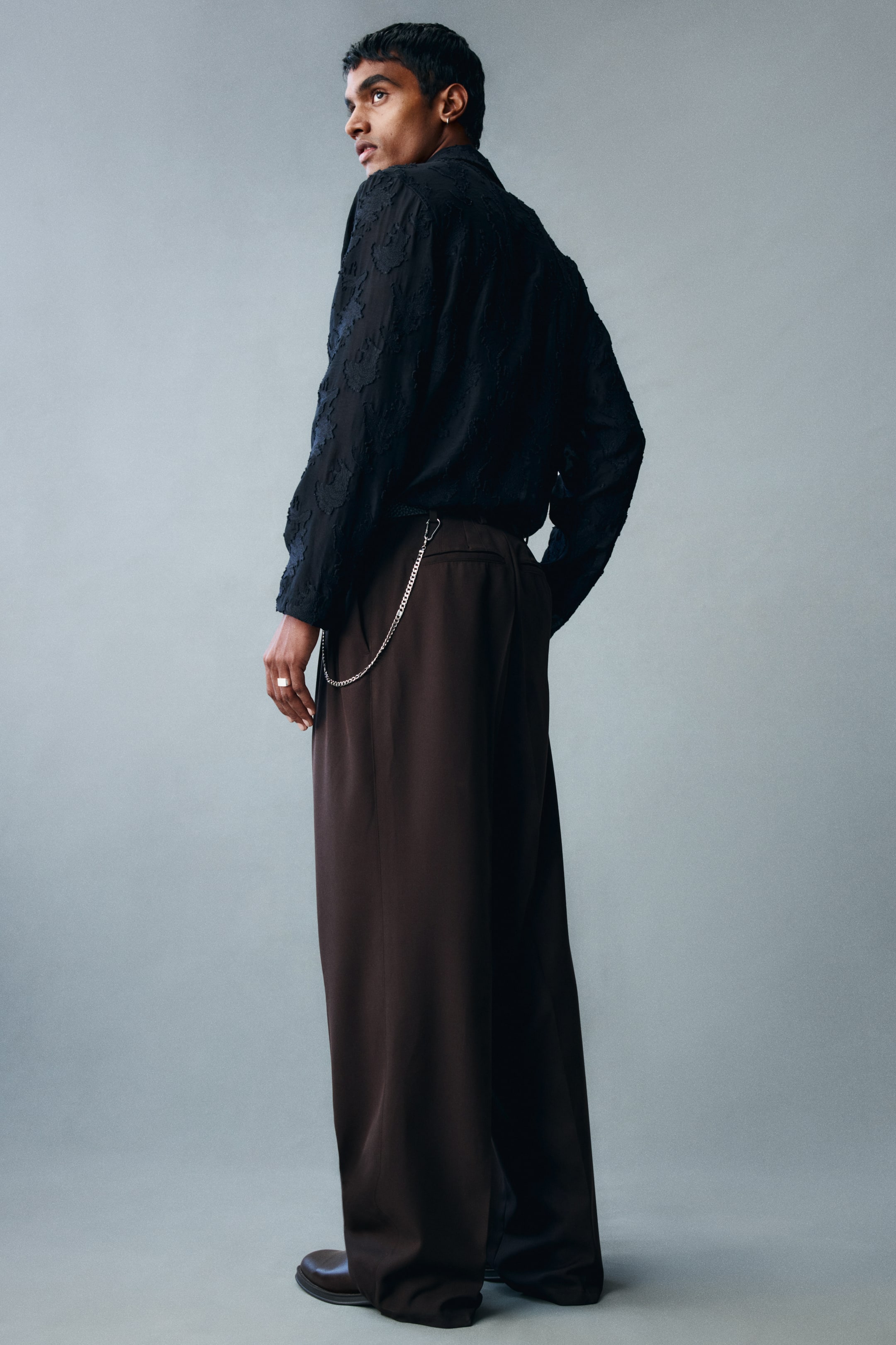 View larger image: The model is wearing tailored pants in dark brown twill, featuring a relaxed, wide-leg silhouette with visible creases running down the back of the legs. The pants are styled with a textured black long-sleeved shirt tucked into the waistband and a silver-toned metal chain draped from a side belt loop.