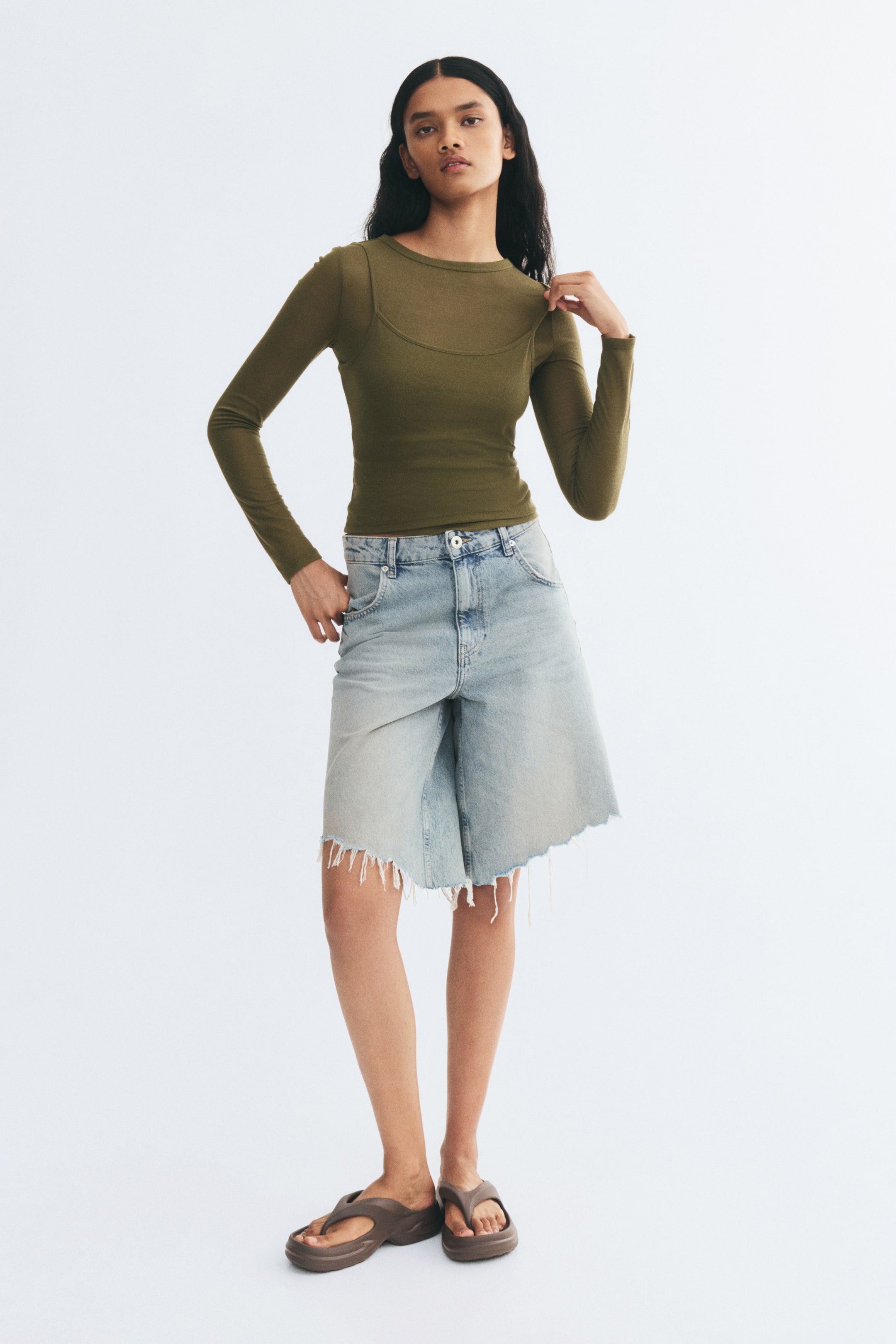 View larger image: A long-sleeve, fitted olive green top in narrow ribbed jersey, featuring a layered design with a scoop-neck outer layer, is worn and lightly adjusted at the shoulder by the model. The top is styled with light-wash, wide-leg denim shorts with frayed hems.
