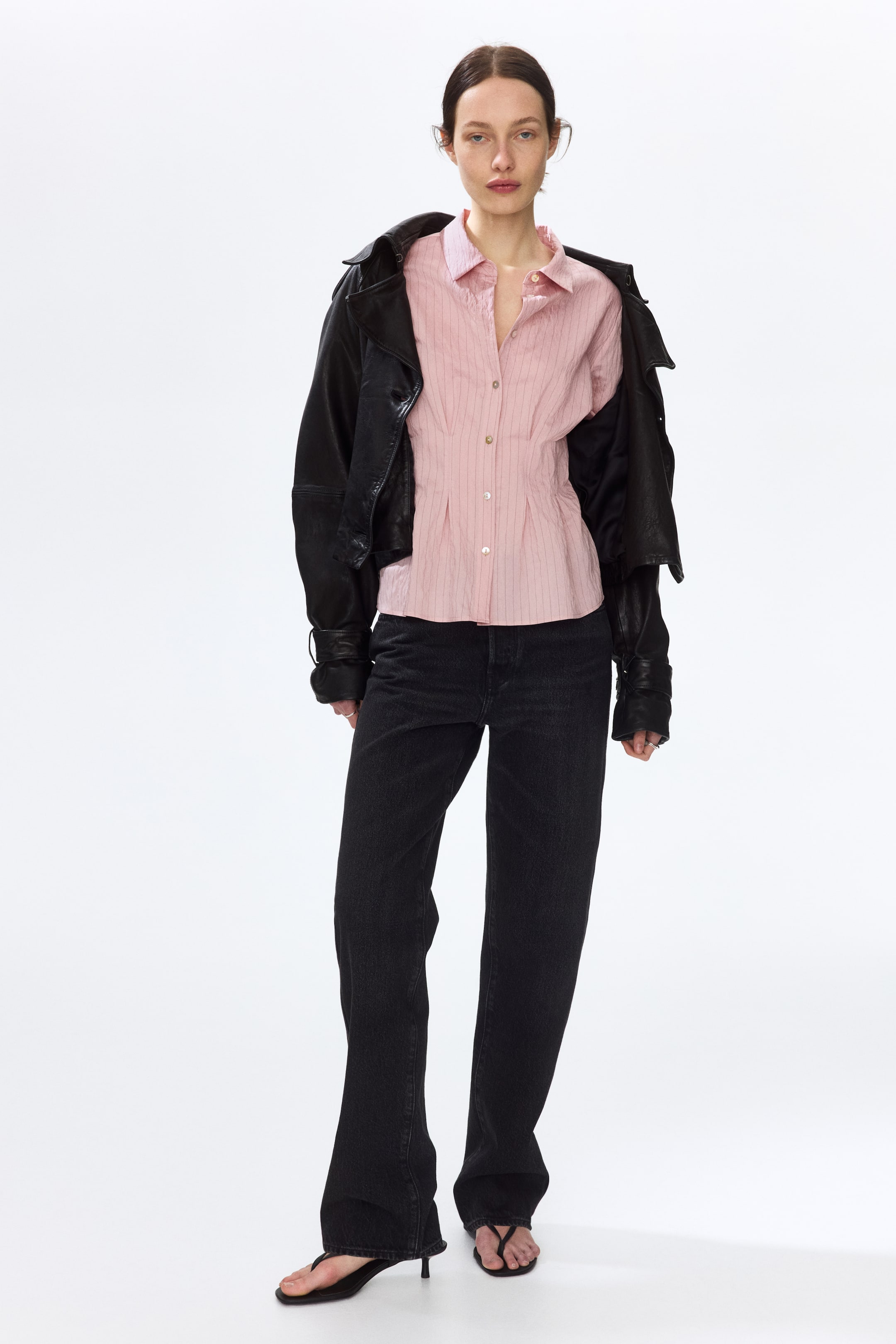 Näytä suurempi kuva: Vaaleanpunainen raidallinen nappipaita, jossa on terävä kaulus, etunapitus ja istuva vyötärö pystysuorilla laskoksilla. Paita on puettuna eteenpäin, tyylitelty mustalla takilla, joka on heitetty olkapäille.