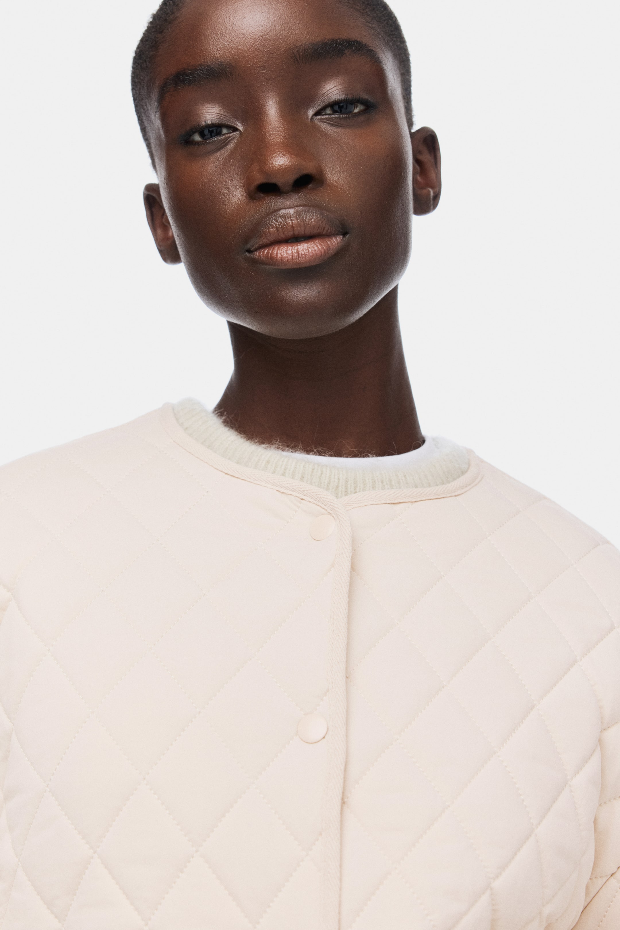 Visualizza immagine più grande: Viene mostrata una giacca trapuntata beige chiaro, caratterizzata da una trapuntatura a rombi, un girocollo e una chiusura frontale con bottoni. Sotto è indossato un morbido collo di maglione a coste, di colore chiaro, visibile all'altezza del collo della giacca.