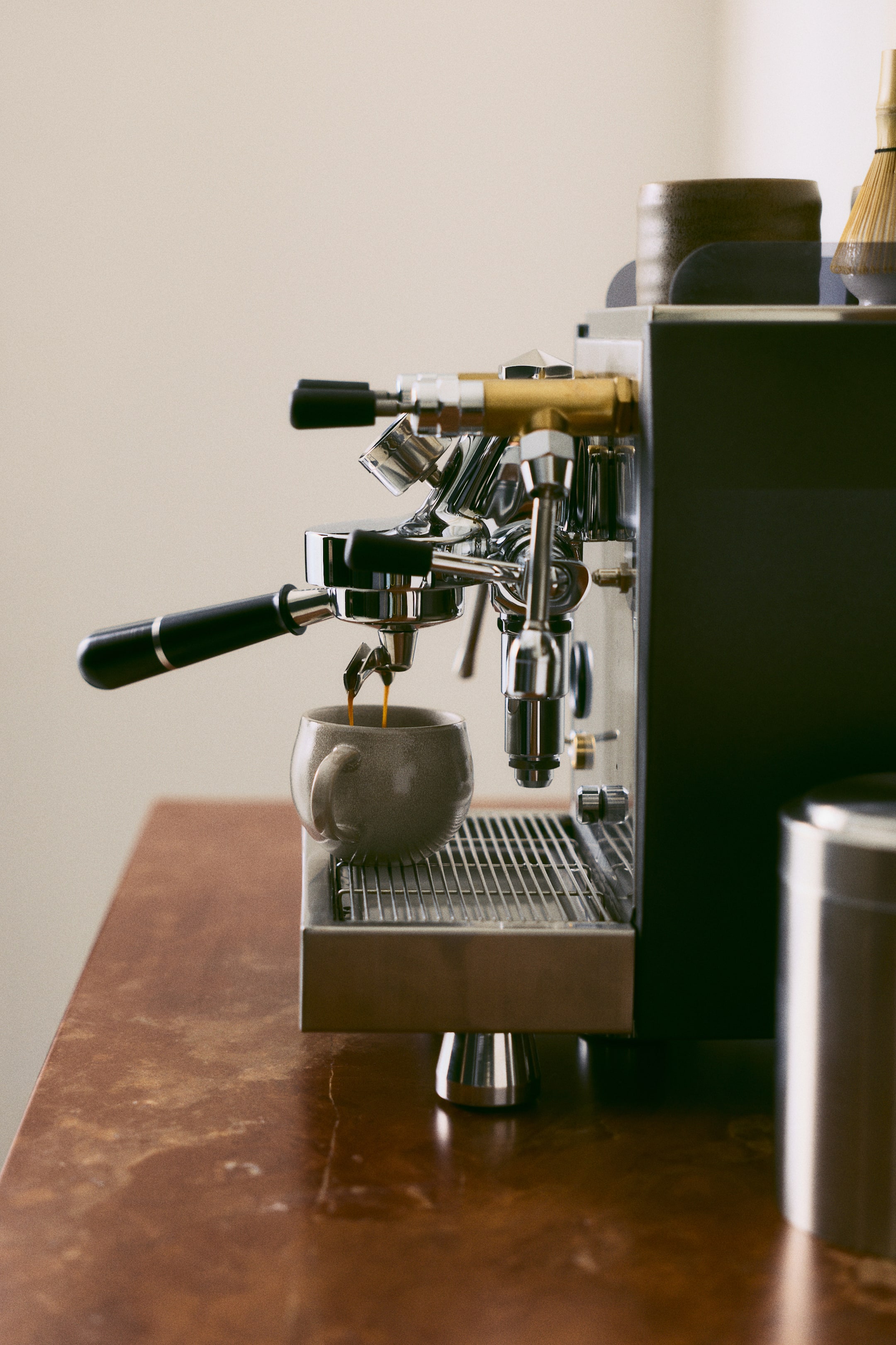 Agrandir l'image: Une tasse en grès présentant une forme arrondie et une petite anse incurvée repose sur une grille en métal tandis que l’espresso y coule goutte à goutte. La tasse arbore un émail réactif brillant, moucheté, dans une teinte gris-beige neutre.