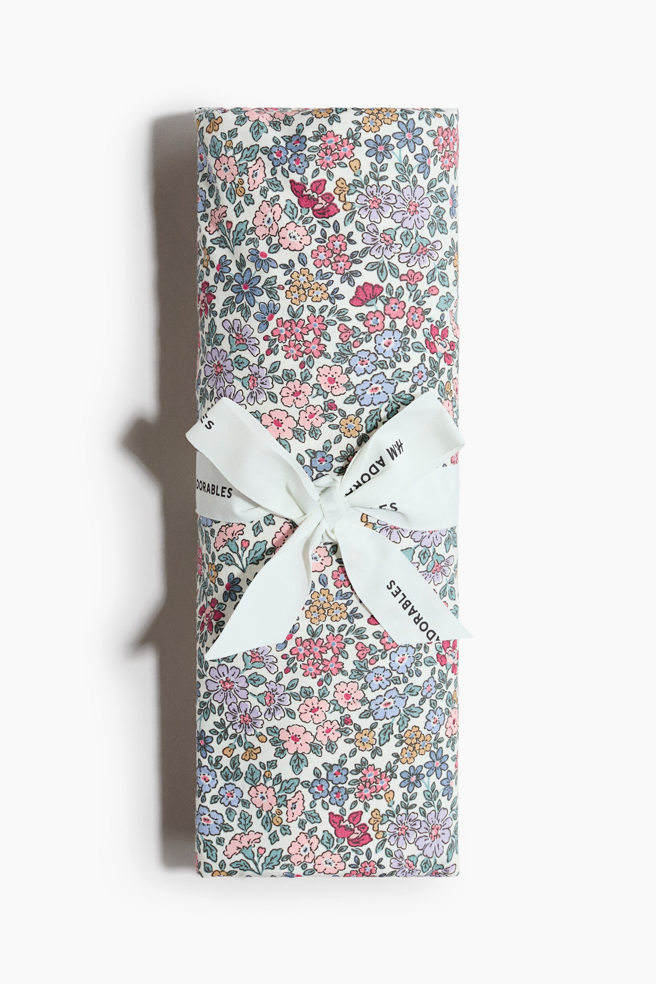 View larger image: A fitted crib sheet is folded into a rectangular package, displaying an all-over floral print in various shades of pink, blue, purple, yellow, and red on a light background. A white ribbon, featuring "H&M ADORABLES" text, is tied in a bow around the middle of the package, visible from the front.