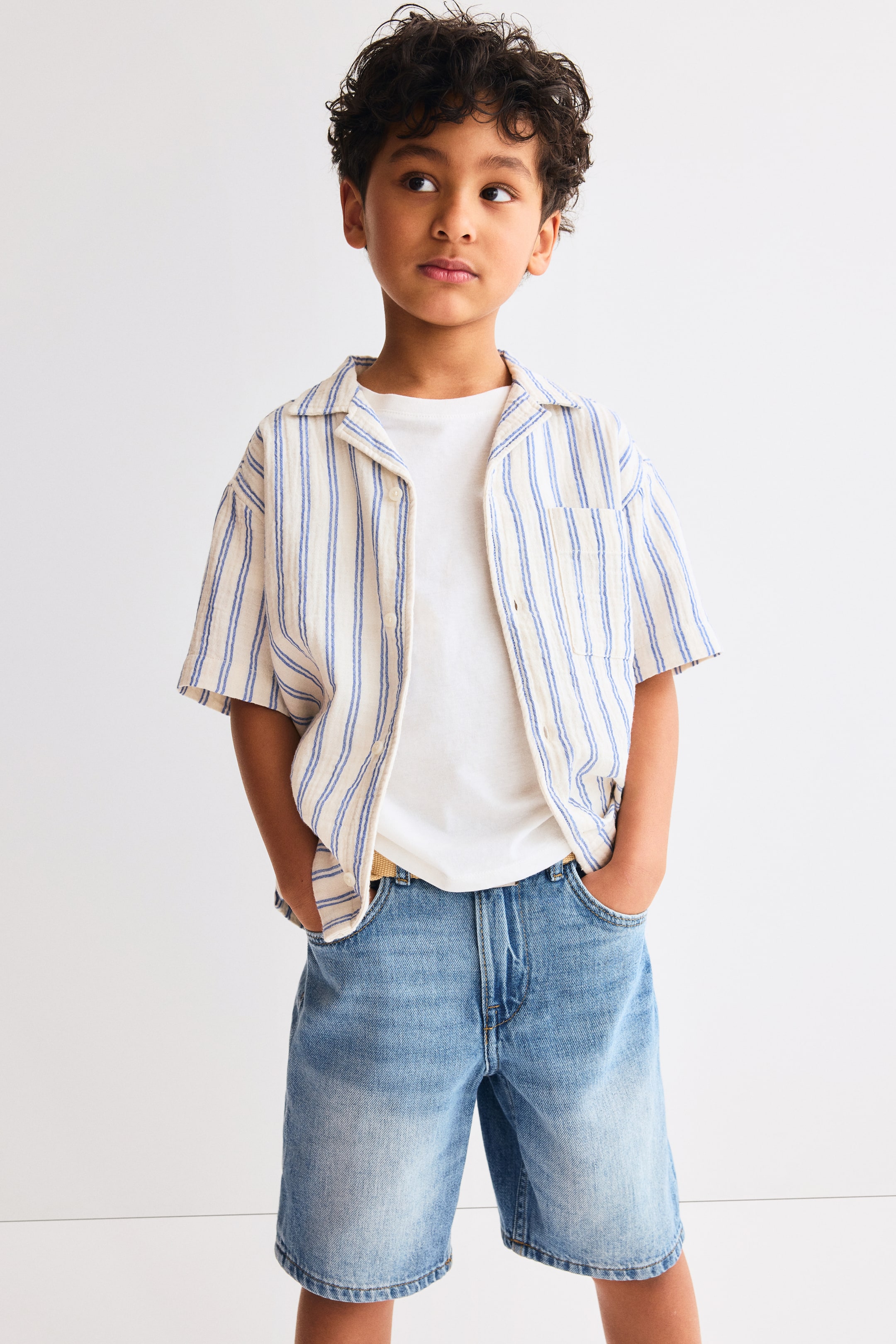 Visualizza immagine più grande: Una camicia a righe beige e blu a maniche corte con colletto alla cubana è indossata aperta sopra una t-shirt bianca a girocollo. Pantaloncini in denim a lavaggio chiaro con tasche anteriori e una cintura chiara visibile si abbinano alle camicie.