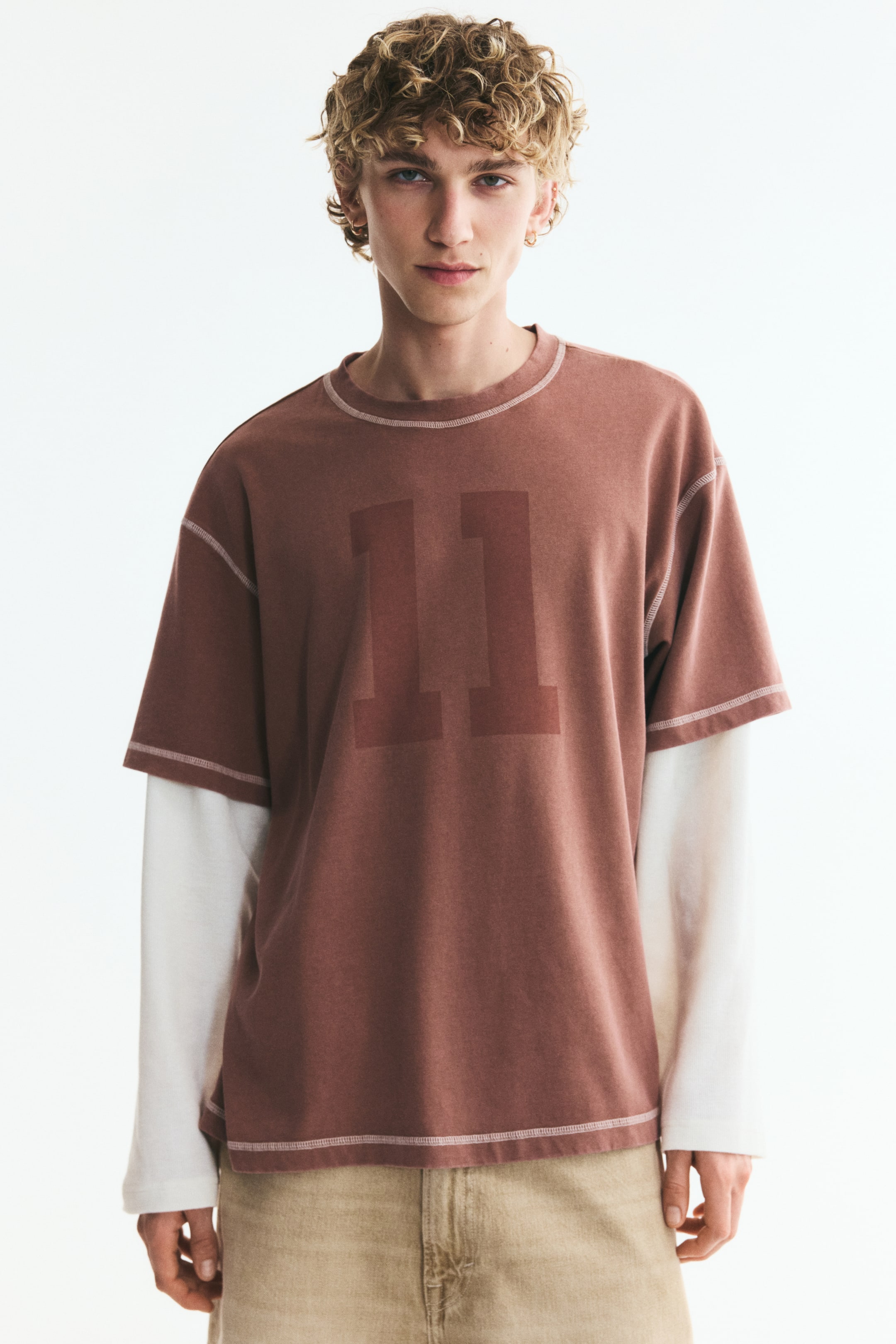 View larger image: A reddish-brown relaxed-fit T-shirt with short sleeves, an "11" print, and visible contrast stitching is worn by the model. It features a rib-trimmed round neck, dropped shoulders, and a straight-cut hem, layered over a white long-sleeved shirt.