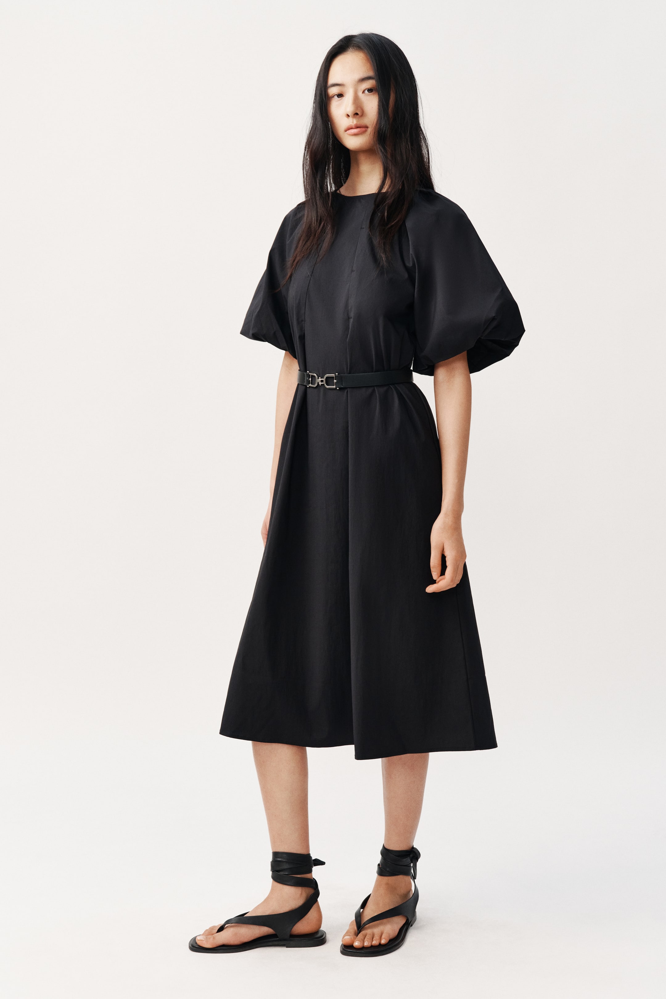 View larger image: This black dress showcases a round neckline and puffed short sleeves. A slim black belt with a silver double-D buckle accentuates the waist, while the A-line skirt elegantly drapes to a midi length.