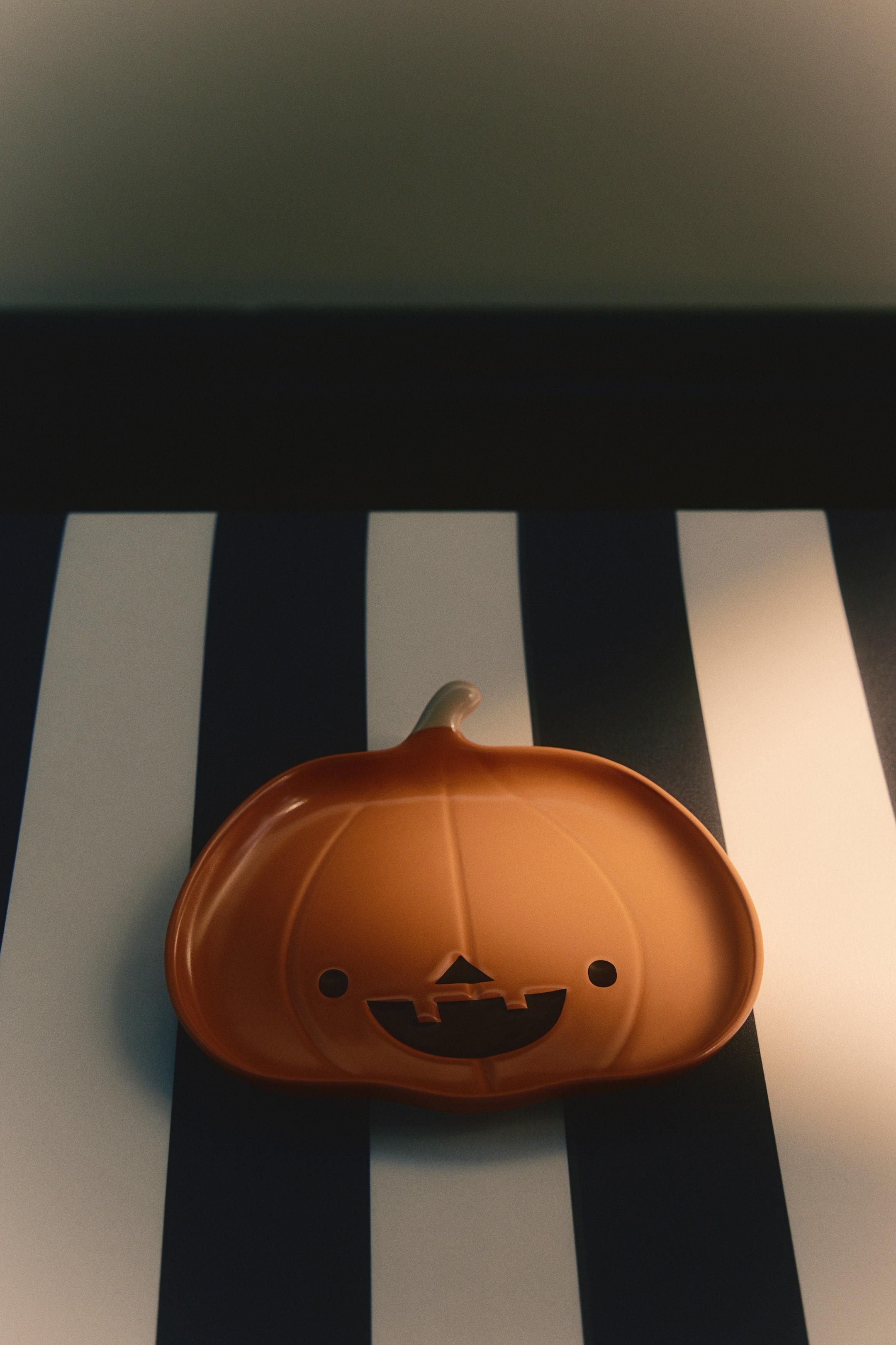 Größeres Bild anzeigen: Ein kürbisförmiger Porzellanteller liegt flach auf und präsentiert seine glänzende orangefarbene Glasur sowie ein stilisiertes schwarzes Jack-o'-Lantern-Gesicht mit runden Augen und einem zahnigen Lächeln. Der Teller verfügt oben über einen kleinen Stiel und dezente vertikale Vertiefungen, die die Segmente des Kürbisses betonen.