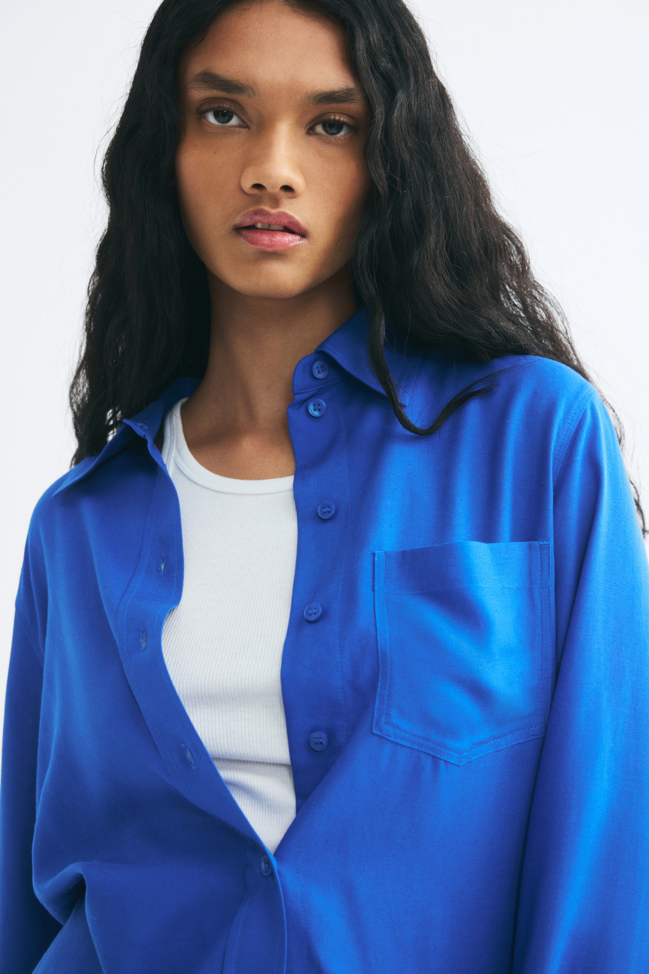 View larger image: A bright blue linen-blend shirt is worn unbuttoned by the model, layered over a white ribbed top. The shirt features a pointed collar, a button-down front, and a single patch pocket on the chest.