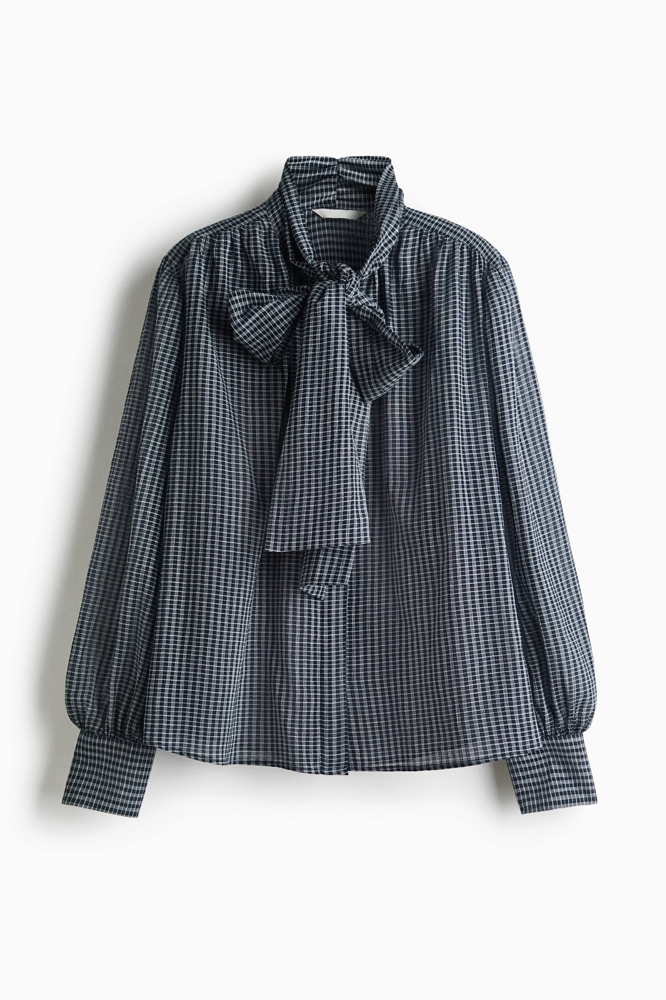 Ver imagen más grande: Una blusa con un pequeño estampado de cuadros en blanco y negro, mostrada de frente, que presenta un cuello alto con lazo anudado en forma de moño y mangas largas y voluminosas fruncidas en los hombros y los puños.