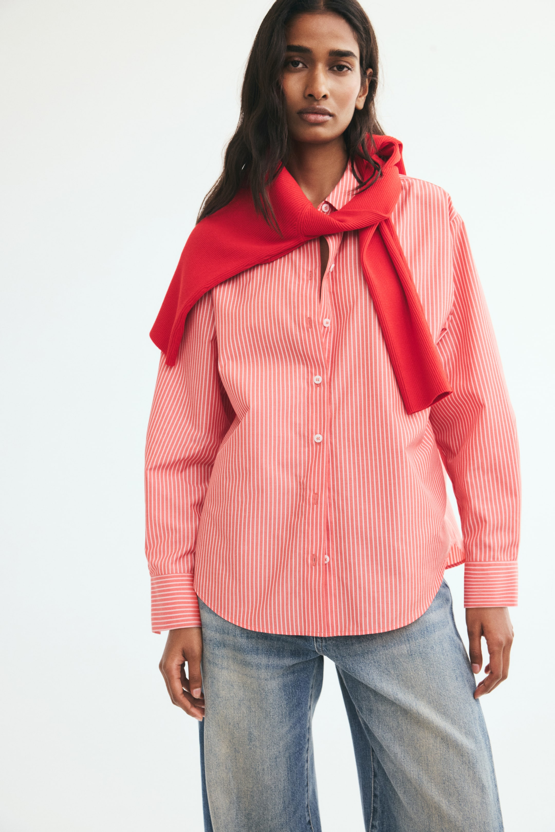 View larger image: A red and white vertically striped woven shirt featuring a collar, button-down front, dropped shoulders, and a rounded hem is worn by the model. The shirt is styled with a solid red sweater draped over the shoulders and paired with light-wash blue jeans.