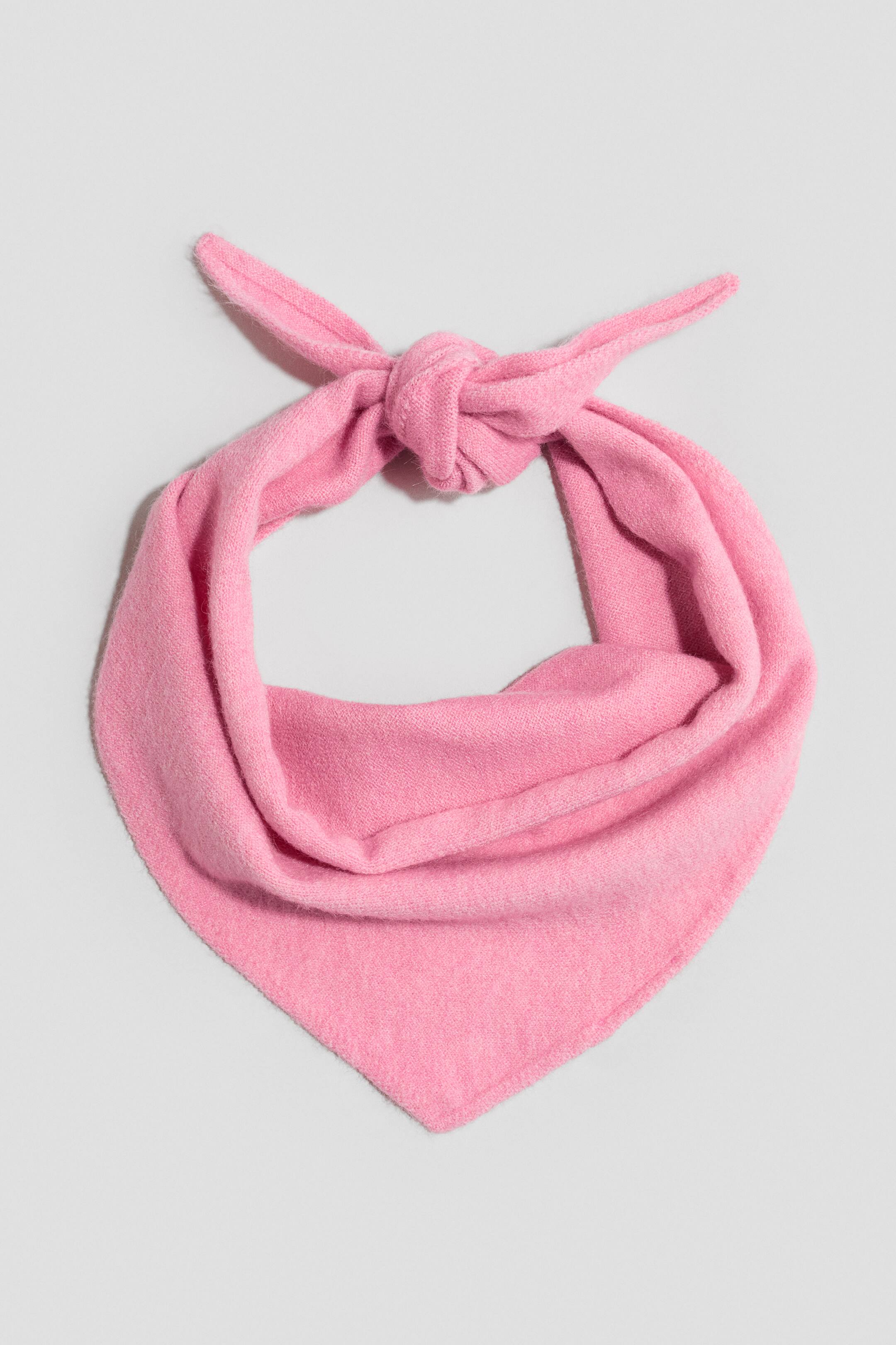 Ampliar la imagen: Un pañuelo de punto rosa está orientado hacia el frente, mostrando una tela suave y texturizada amarrada en un nudo en la parte superior. La bufanda cae hacia abajo para formar un lazo circular que se afina hasta una punta triangular.