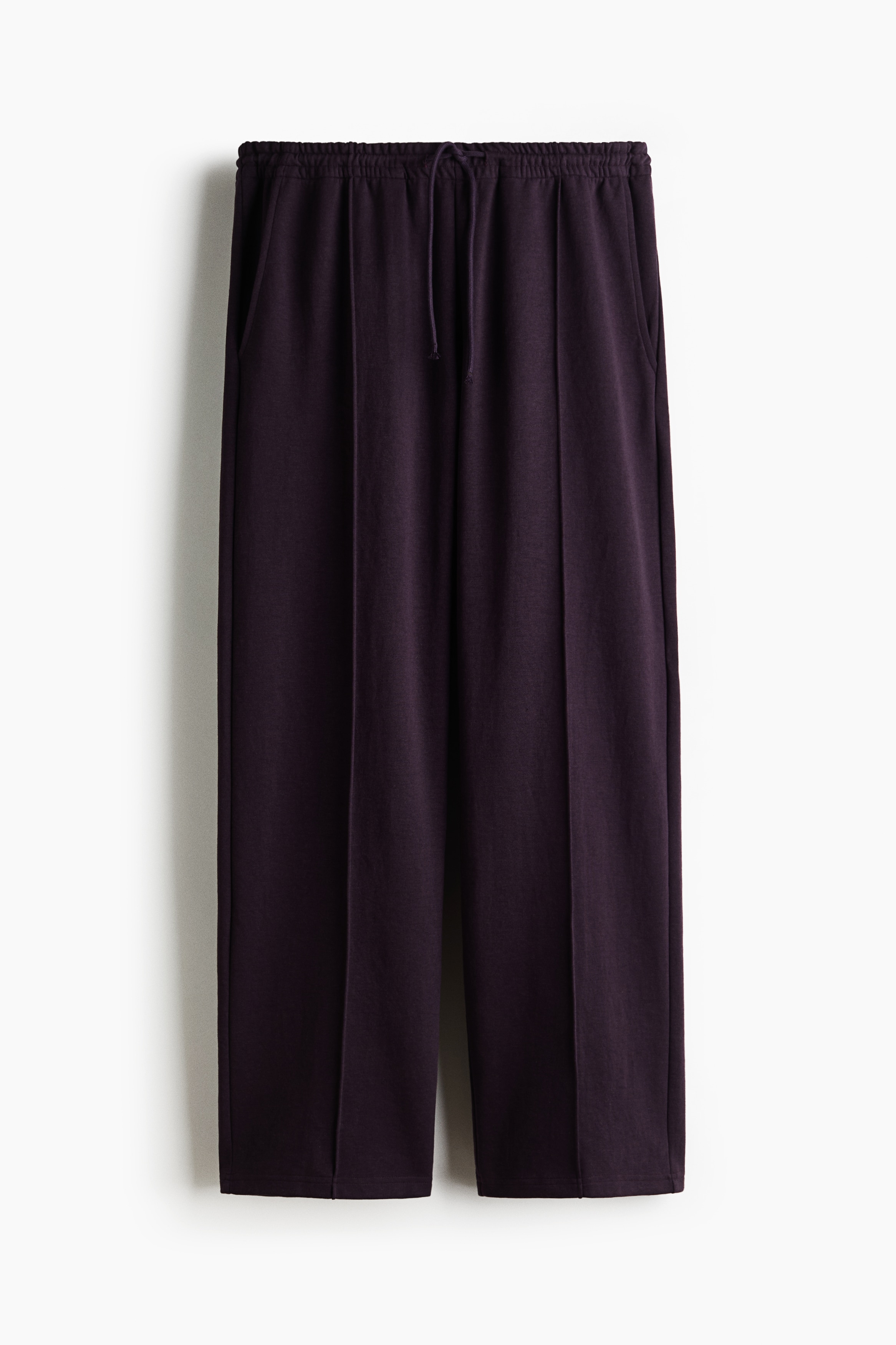 Dark purple relaxed-fit joggers with straight legs and stitched front creases are worn by the model, who is standing with hands in the side pockets. The joggers are paired with a white short-sleeved T-shirt and black sneakers.