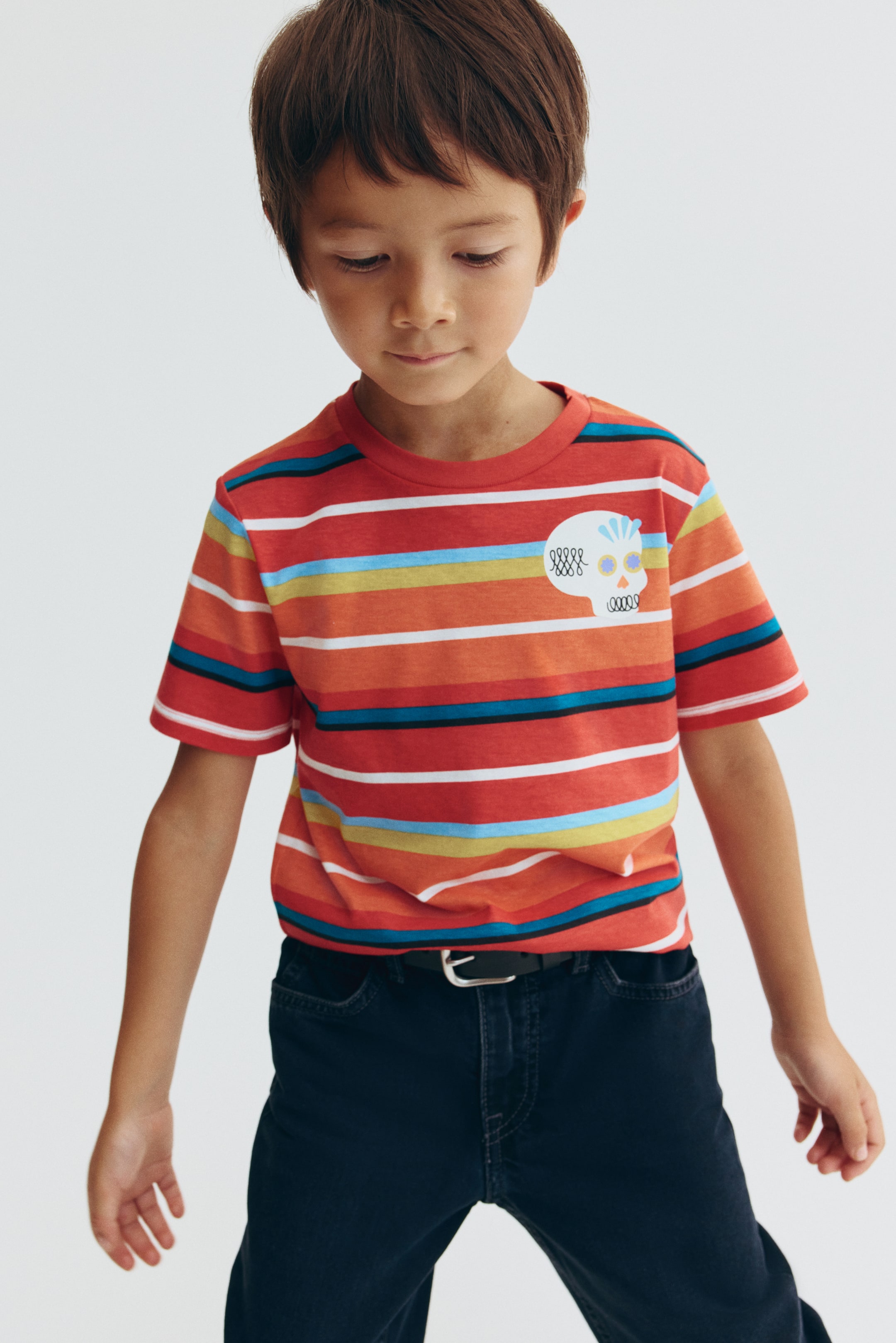 View larger image: A striped cotton jersey tee is shown from the front, featuring multicolored horizontal stripes in red, orange, blue, yellow, and white, with a small decorative skull print on the left chest. The tee is designed with a round, rib-trimmed neckline and dropped shoulders, and is shown tucked into dark-colored pants.