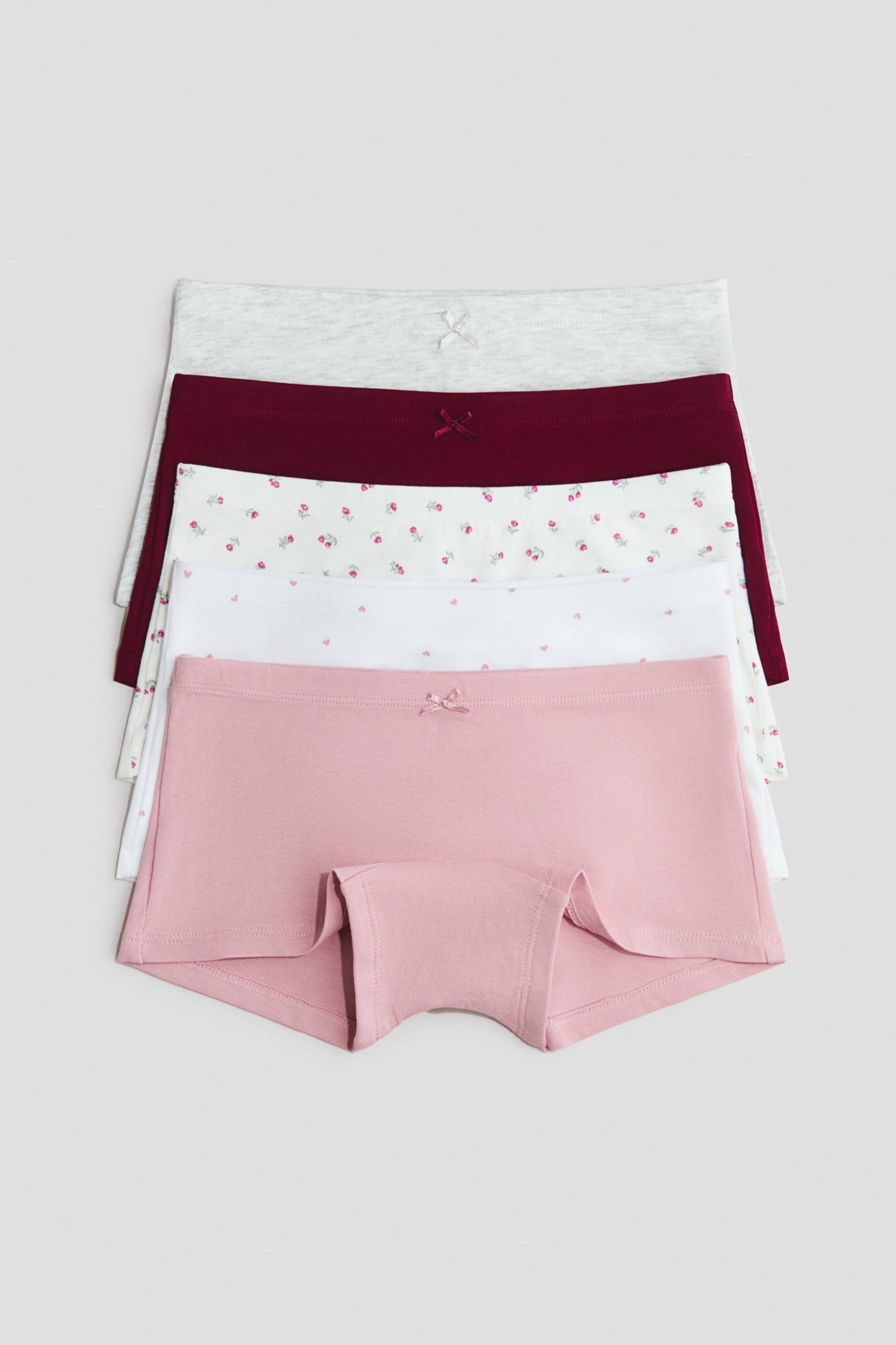 Größeres Bild ansehen: Ein Fünferpack Boyleg-Slips aus Baumwolljersey liegt von vorne flach in gestapelter, versetzter Anordnung und zeigt den Boyleg-Schnitt sowie die elastischen Bündchen. Das Set umfasst im Vordergrund ein einfarbiges Paar in Staubrosa mit einer kleinen Zierschleife, das über Paaren mit pinkfarbenem Herzprint, Blumenmuster, einfarbigem Burgunderrot und hell meliertem Grau liegt.