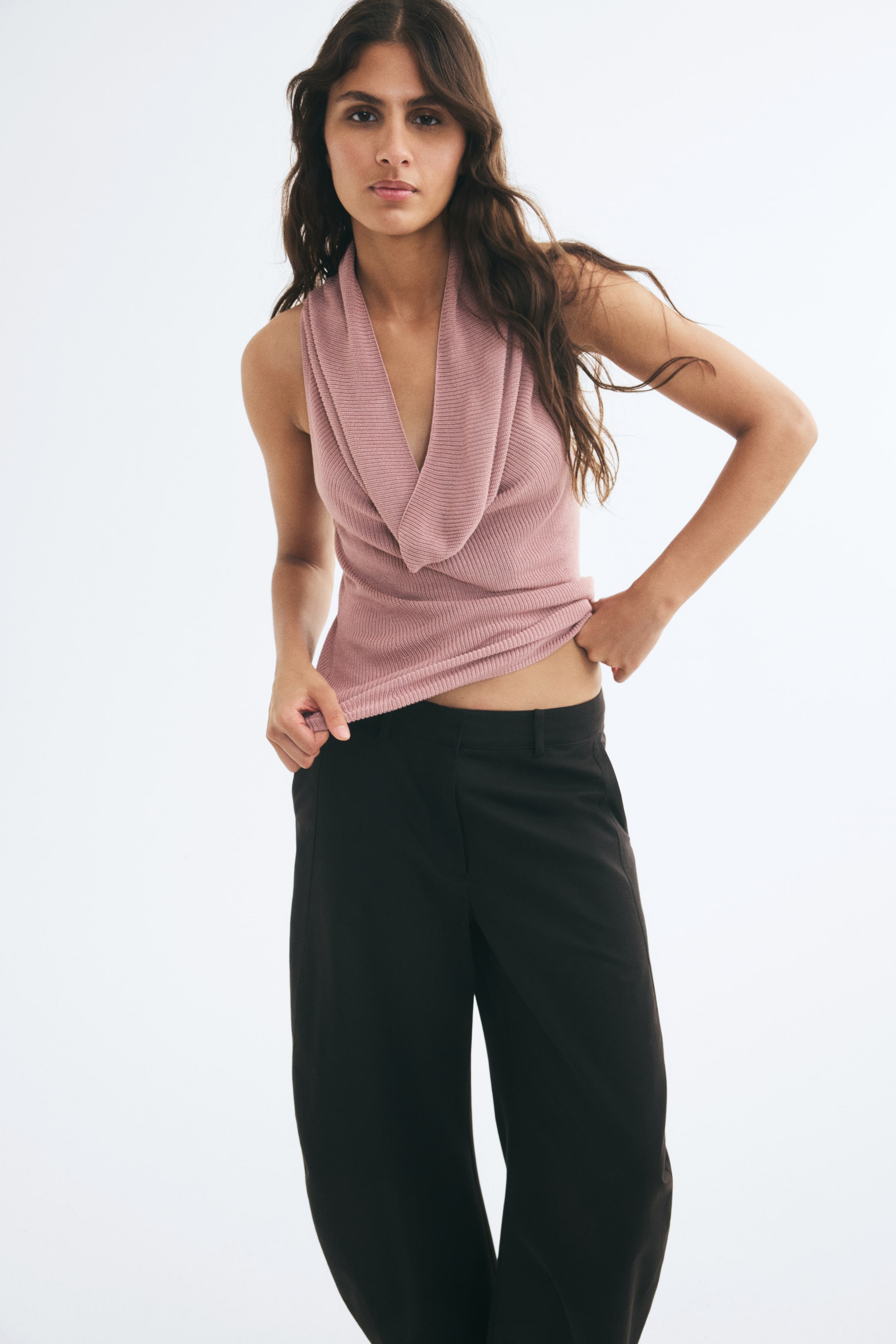 Ampliar la imagen: Una blusa rosa de punto canalé sin mangas, con un profundo escote drapeado, es llevada por la modelo, quien sostiene ligeramente levantado el dobladillo recto a la altura de la cintura. La blusa se combina con pantalones negros de pierna ancha.