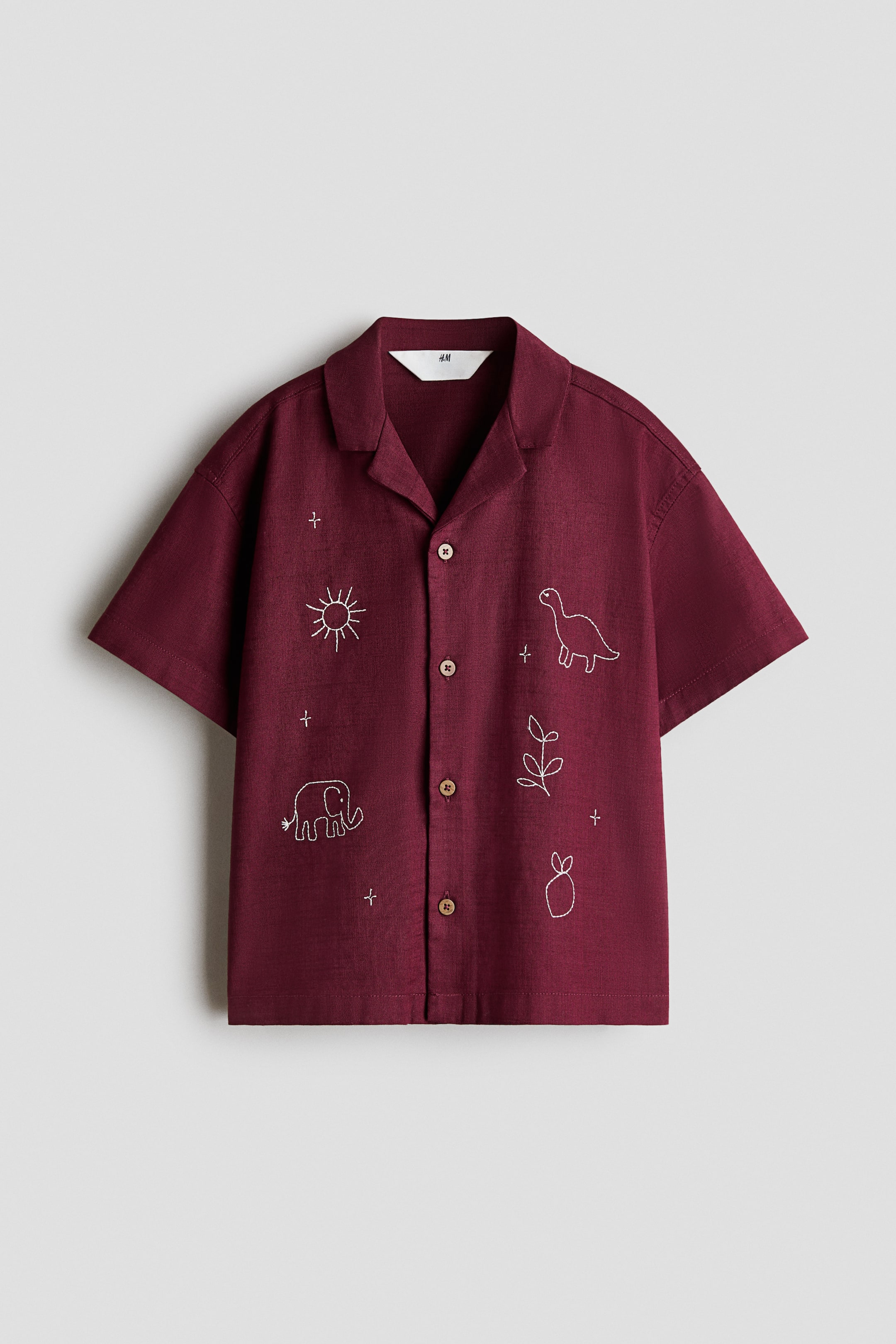 View larger image: A burgundy short-sleeve shirt is shown from the front, featuring a resort collar, dropped shoulders, and a button-down front with four light-brown buttons. The shirt displays white embroidered line drawings of an elephant, a dinosaur, a sun, stars, a leafy branch, and a pineapple scattered across both front panels.