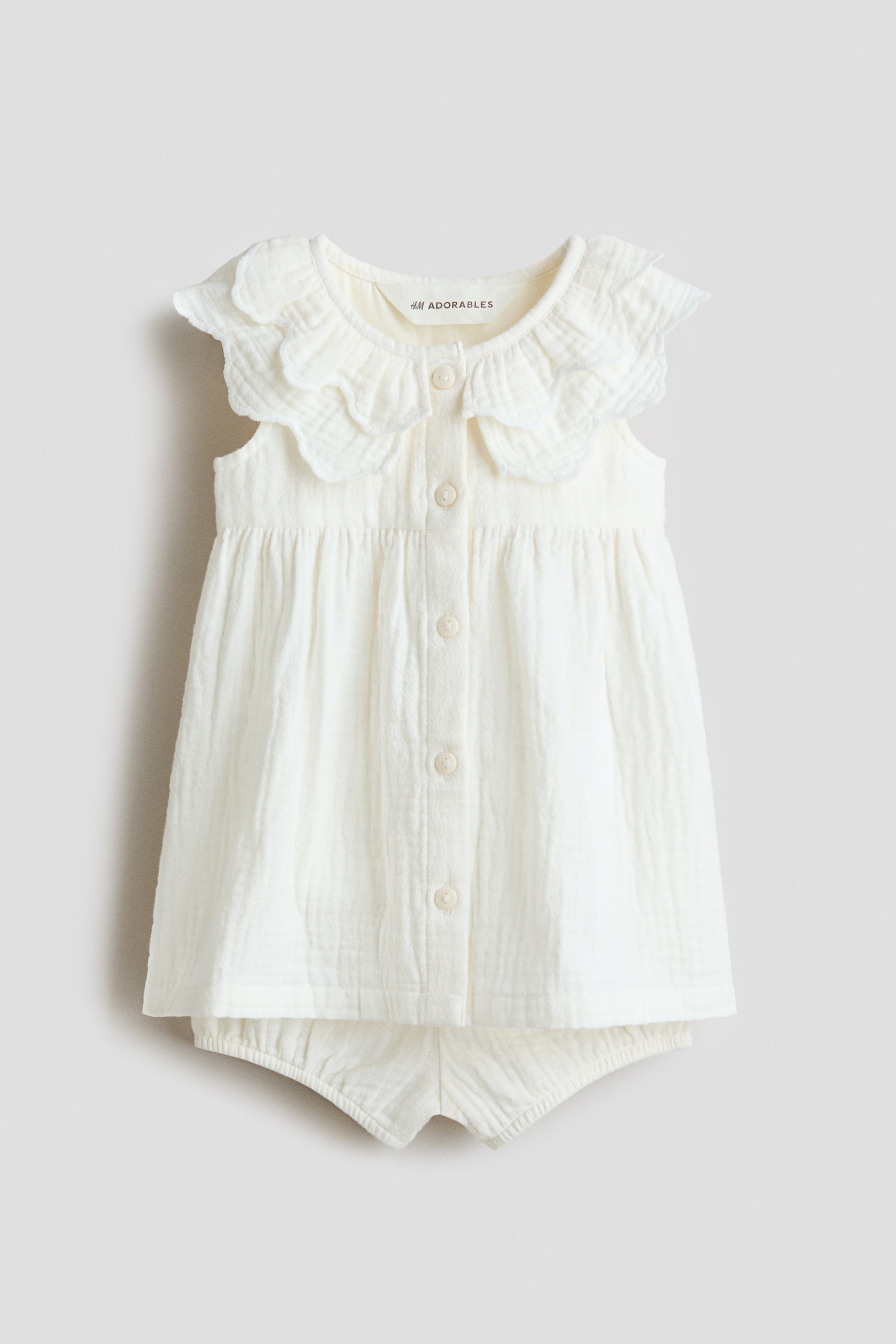 Voir une image plus grande: Un ensemble pour bébé de couleur crème, confectionné en popeline douce, est présenté de face, comprenant une robe sans manches et des bloomers assortis. La robe arbore un col à volants festonnés à double épaisseur au niveau de l'encolure ronde, avec des boutons parcourant le devant et une couture froncée au-dessus de la taille créant une jupe évasée, tandis que les bloomers assortis sont visibles en dessous, affichant des ouvertures de jambes élastiquées.