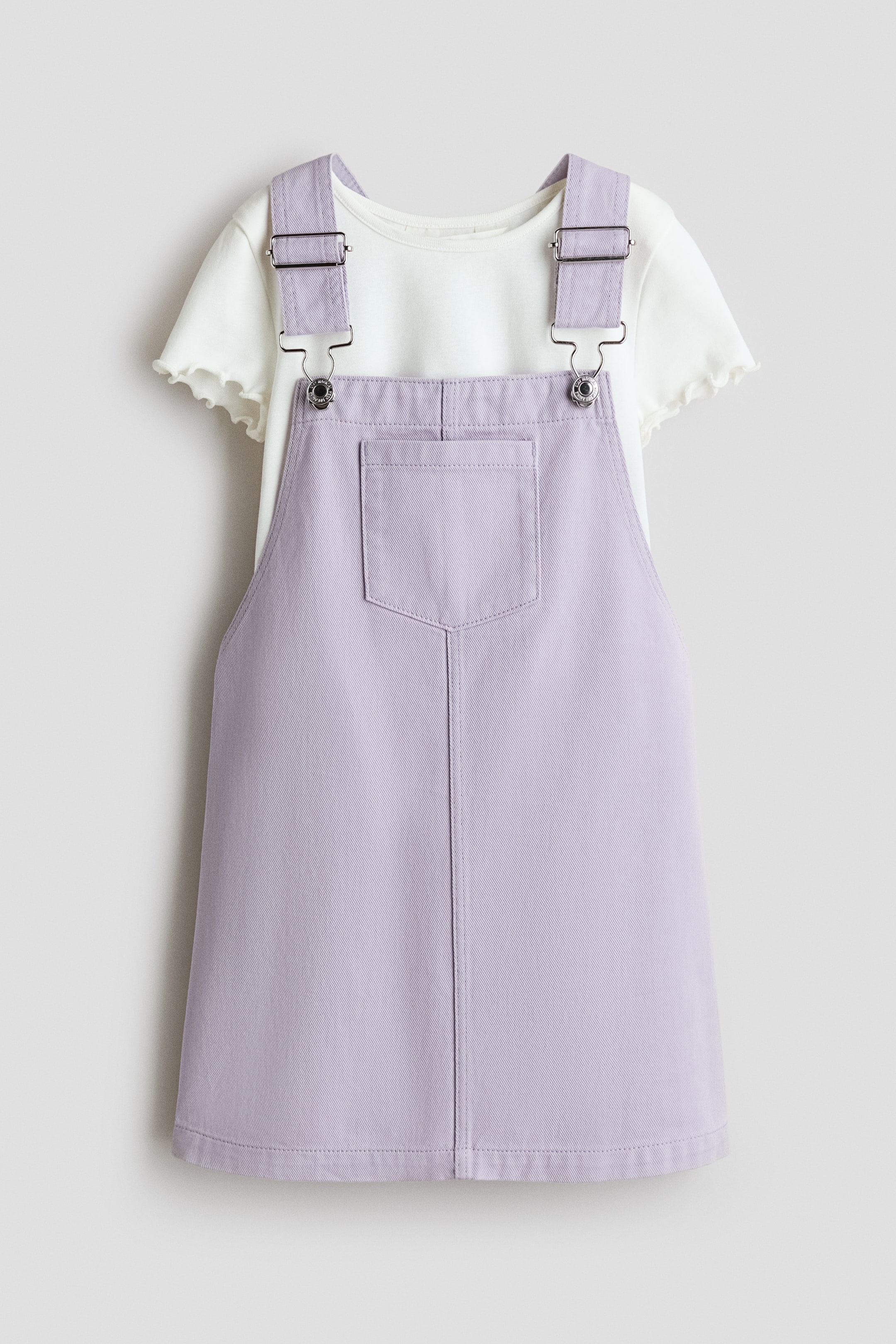View larger image: The set features a light purple overall dress shown from the front, with adjustable shoulder straps finished with metal buckles and a single rectangular chest pocket. The dress is layered over an off-white T-shirt with a narrow neckline trim and serged, wavy edges at the short sleeve cuffs.