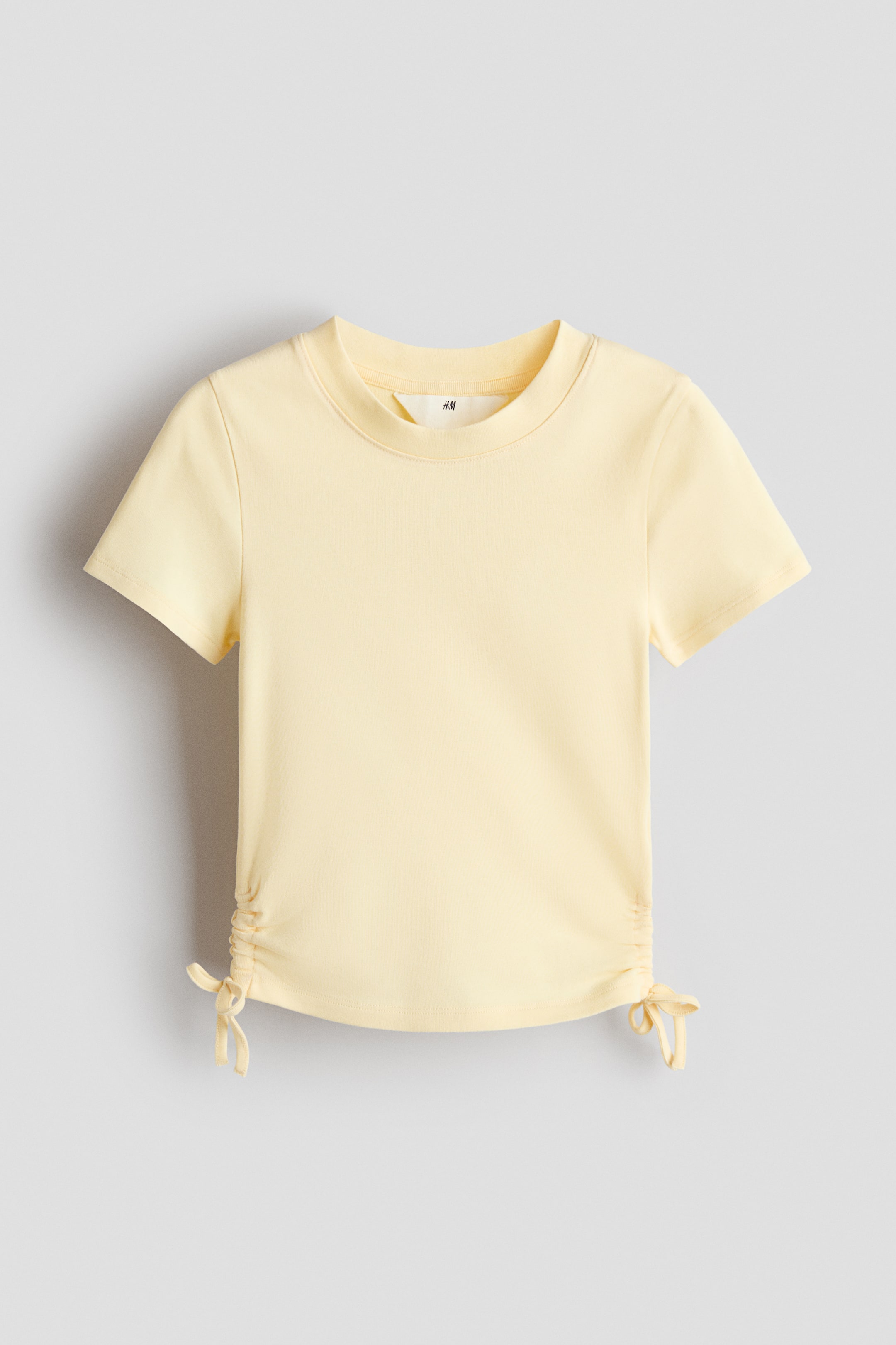 View larger image: A light-yellow, short-sleeved top faces forward and features a crew neck. It has ruched sides with ties at the hem, and an "H&M" label is visible inside the neckline.