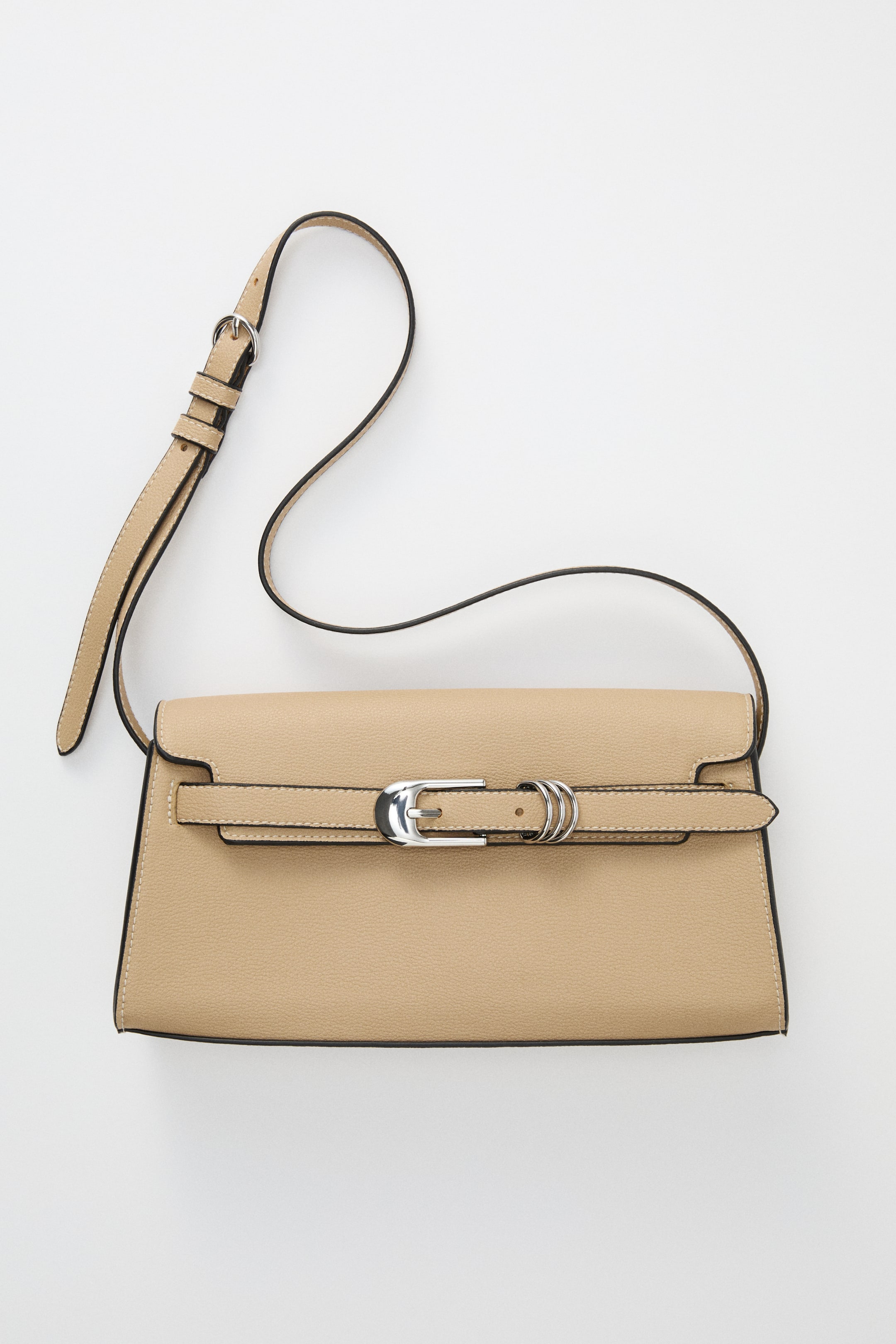 View larger image: A beige grained shoulder bag is presented from the front, showcasing a flap closure that is detailed with a horizontal strap featuring a silver-toned metal buckle and two D-rings. An adjustable shoulder strap with a metal buckle is extending from the bag's top.