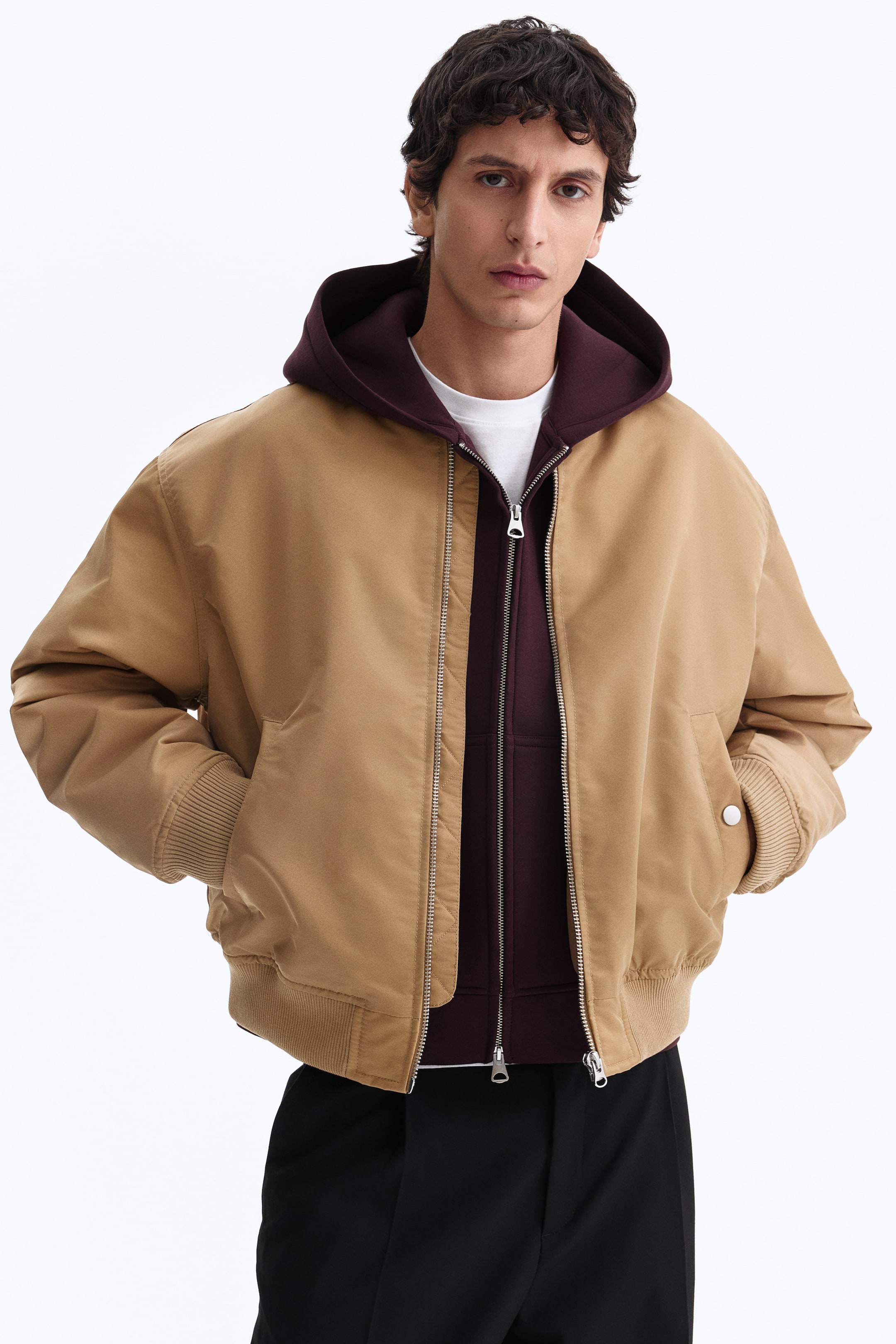 View larger image: A tan bomber jacket is shown from the front, slightly angled to the right, featuring a silver-toned zipper closure, ribbed cuffs, and a ribbed hem. It is layered over a maroon zip-up hoodie and a white t-shirt.
