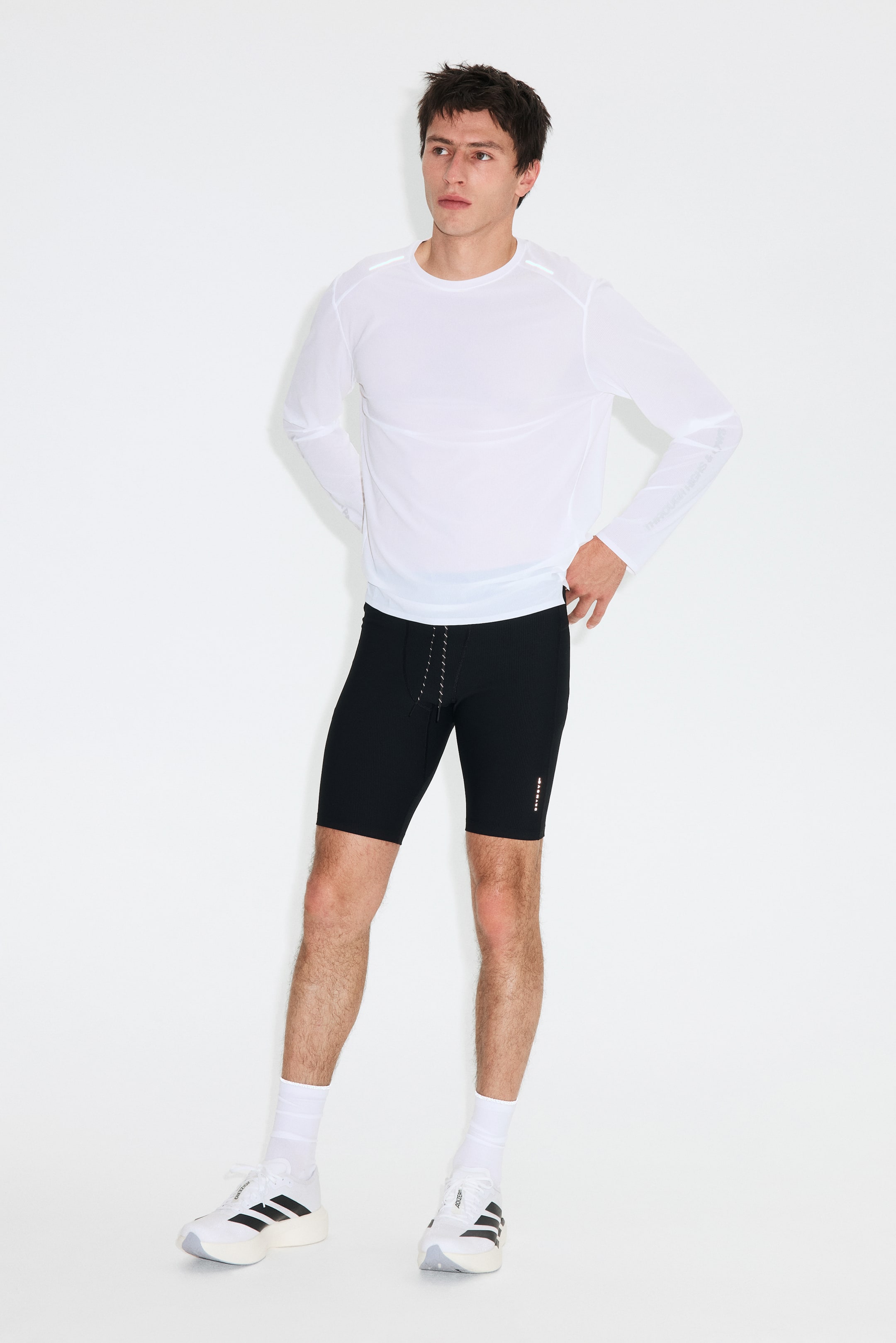 View larger image: The athletic ensemble comprises a white long-sleeve top paired with black form-fitting shorts, styled facing forward with a slight right angle. The top features reflective elements on the shoulders and text on the left forearm, highlighting its snug fit. The shorts showcase a drawstring waist and reflective details on the left thigh. Completing the look are white mid-calf socks and white athletic shoes adorned with black stripes.