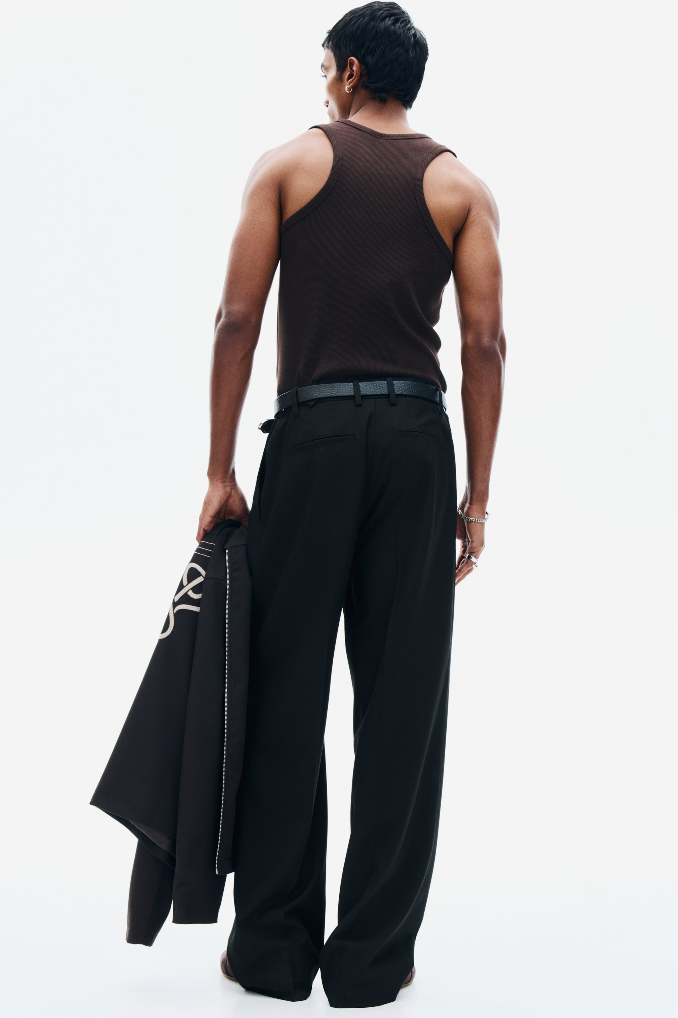 View larger image: The model is wearing tailored black twill trousers, featuring a relaxed fit, straight legs, and pressed back creases. The trousers are styled with a black belt, and the model is holding a dark jacket in one hand.