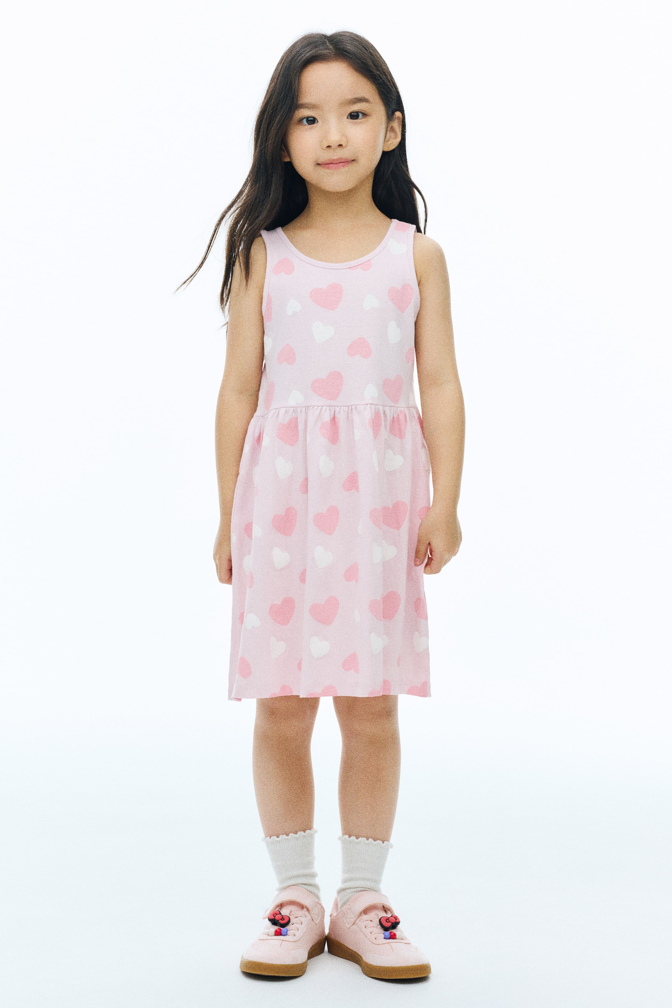View larger image: A sleeveless pink jersey dress with an all-over print of white and pink hearts is worn by the model. The dress features a round neckline, a gathered seam at the waist, and a flared skirt.